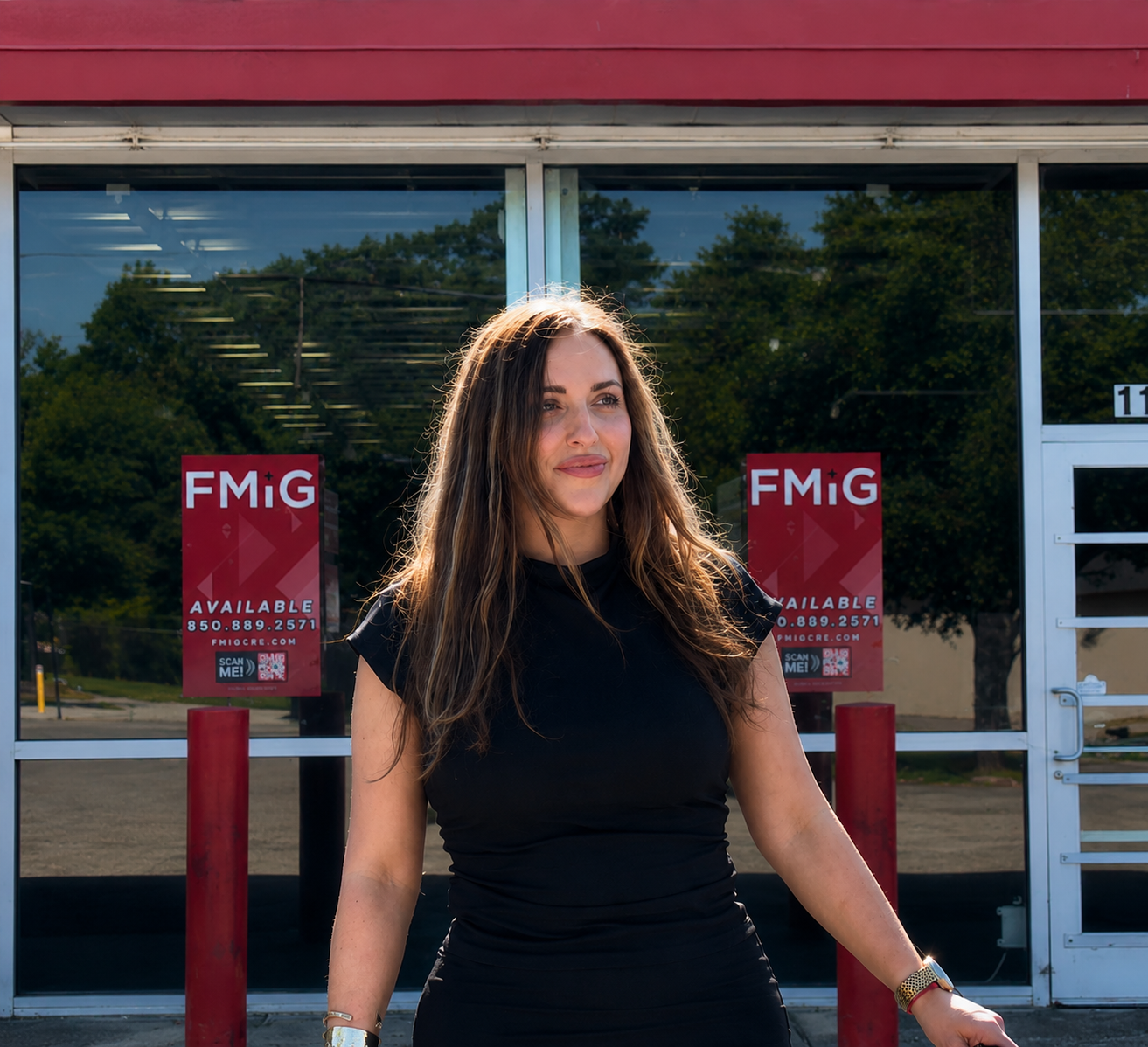 A woman with long brown hair in a black shirt standing outside a glass storefront with red and white signs that say 'FM+G' and 'Available' with contact information.