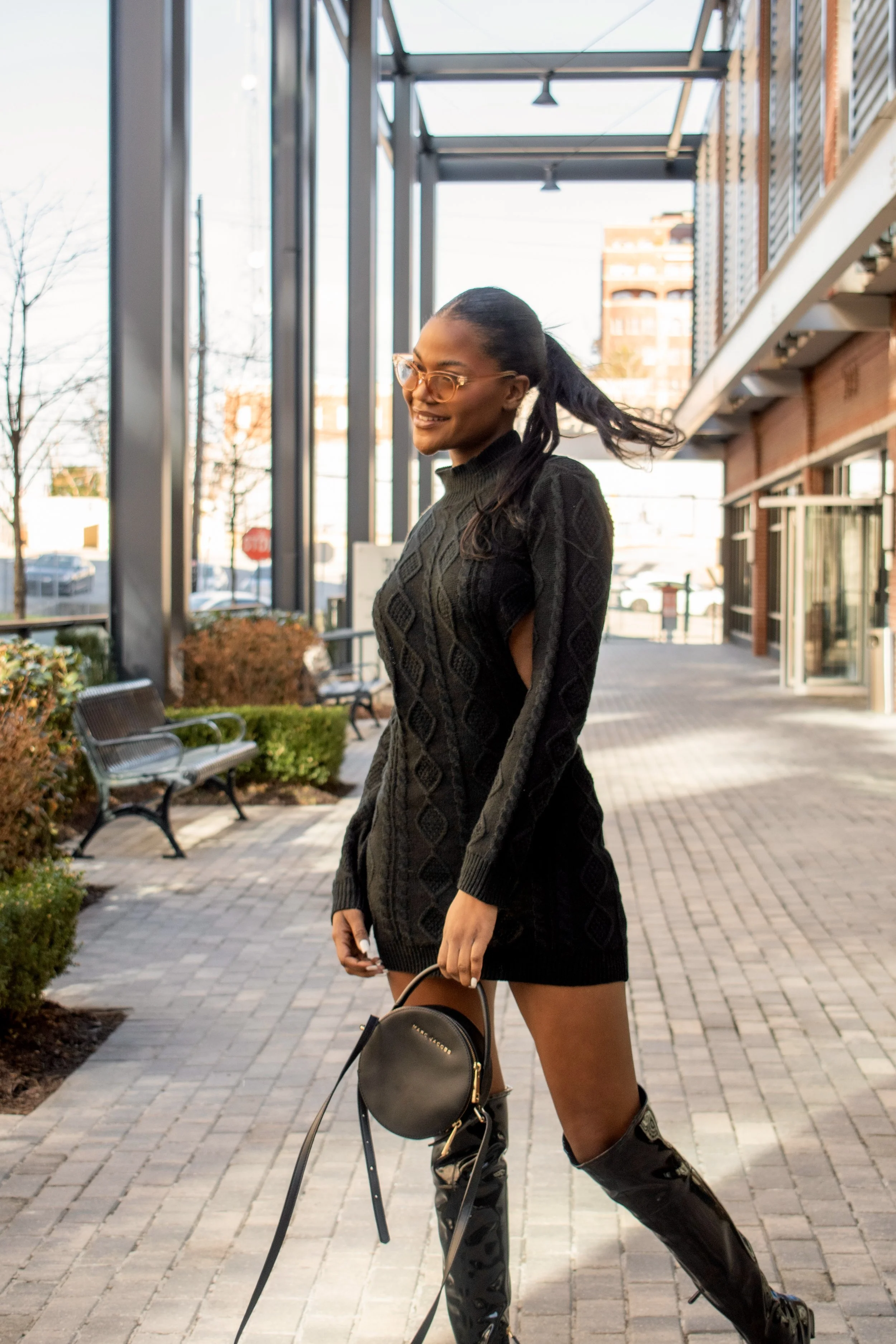 Fashionable woman in black sweater dress and thigh-high boots holding a handbag, standing on a city sidewalk.