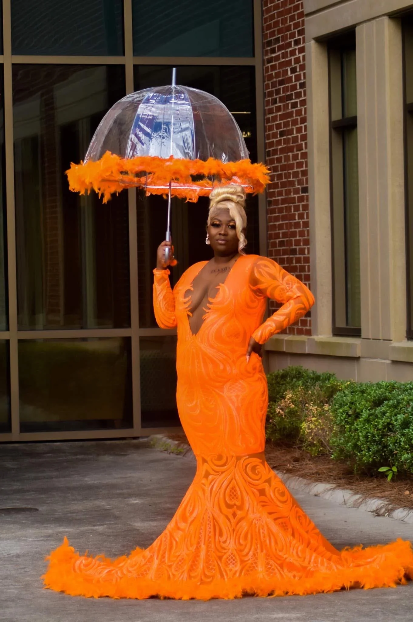 Person in an orange gown holding a clear umbrella with orange trim, standing in front of a windowed building.