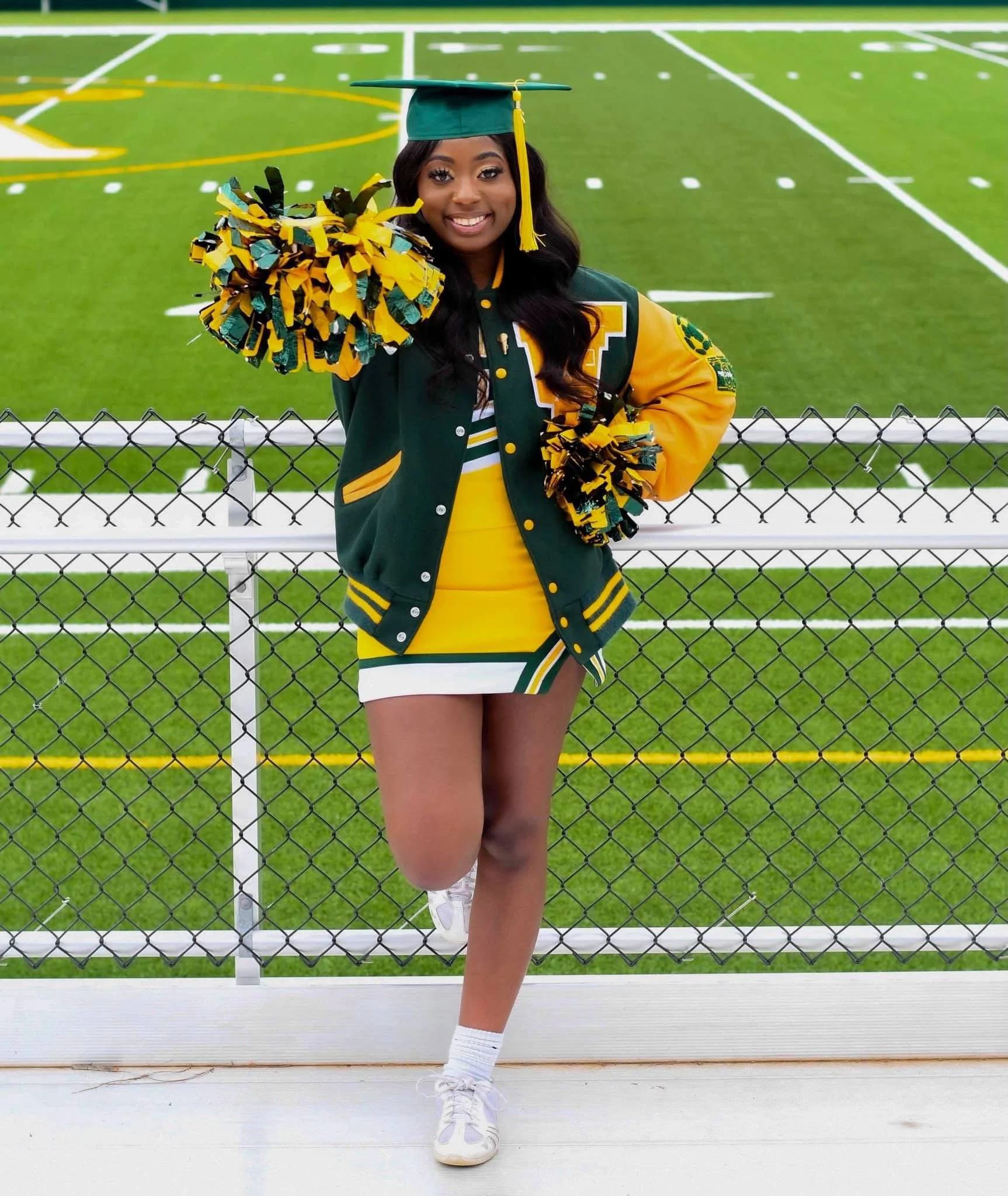 A cheerleader in a green and yellow uniform poses with pom-poms on a sports field. She is wearing a green graduation cap and a varsity jacket, smiling at the camera.