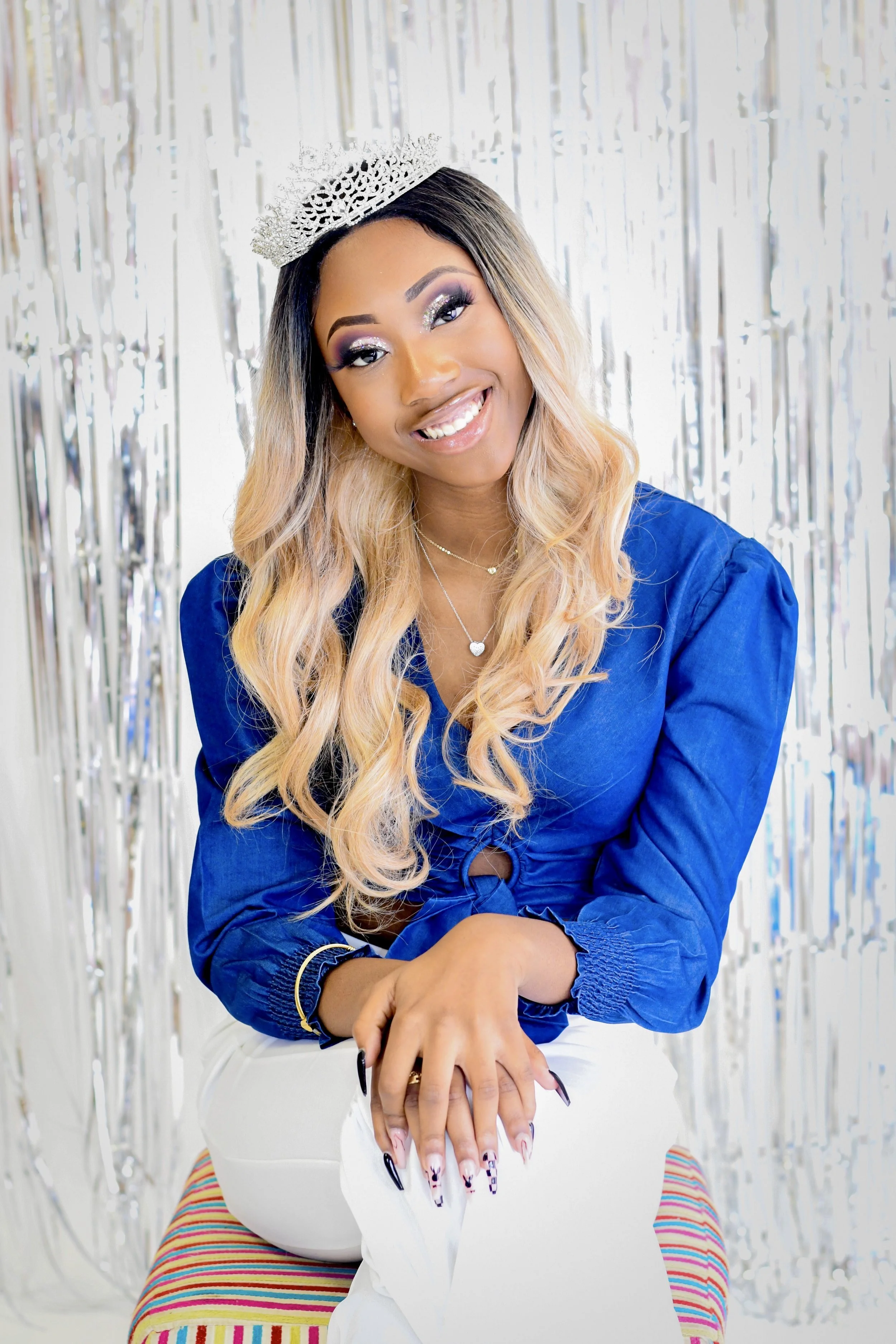 Woman wearing a blue blouse with a crown, smiling and sitting in front of a silver fringe background.