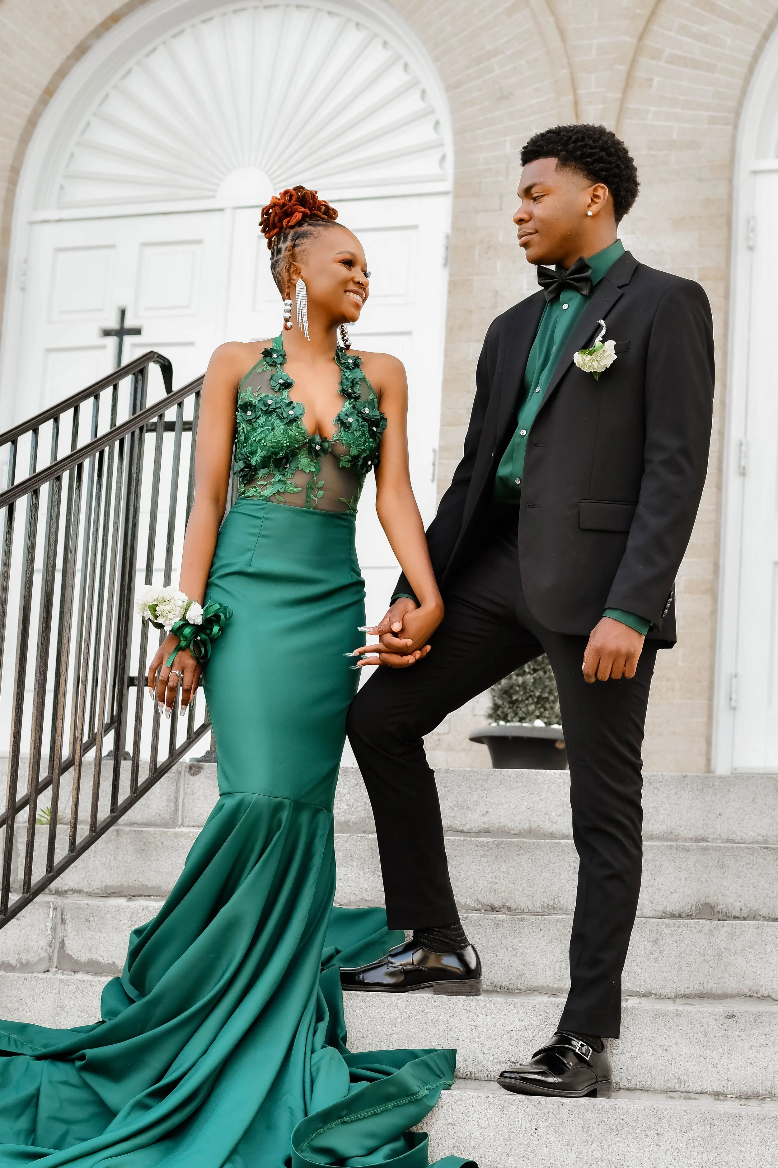 A couple in formal attire stands on steps in front of a building. The woman is wearing a green floral gown with a long train, and the man is in a black suit and bow tie with a green shirt. They are smiling and holding hands.