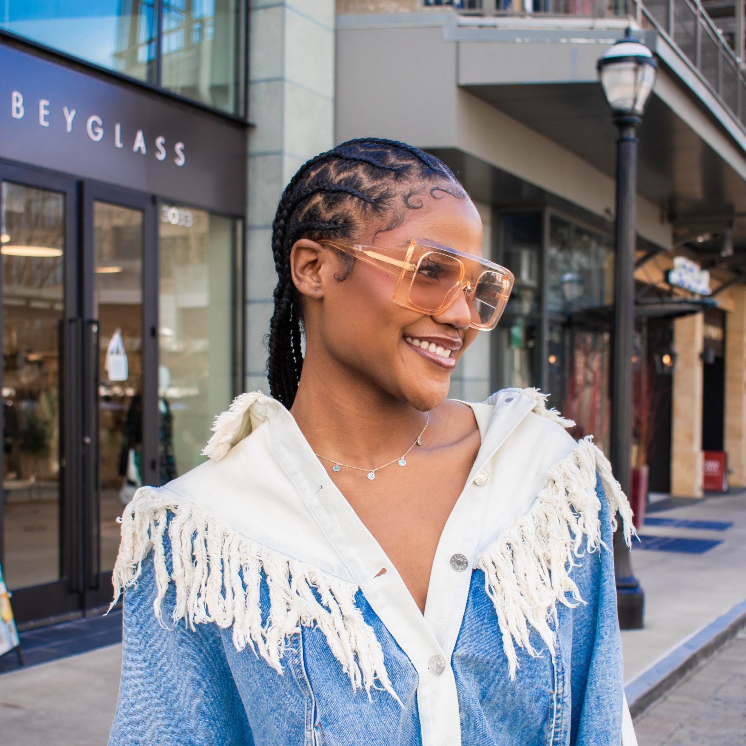 Fashionable woman wearing large sunglasses and denim jacket with fringe detail, stands smiling outside a store with a BEYGLASS sign.
