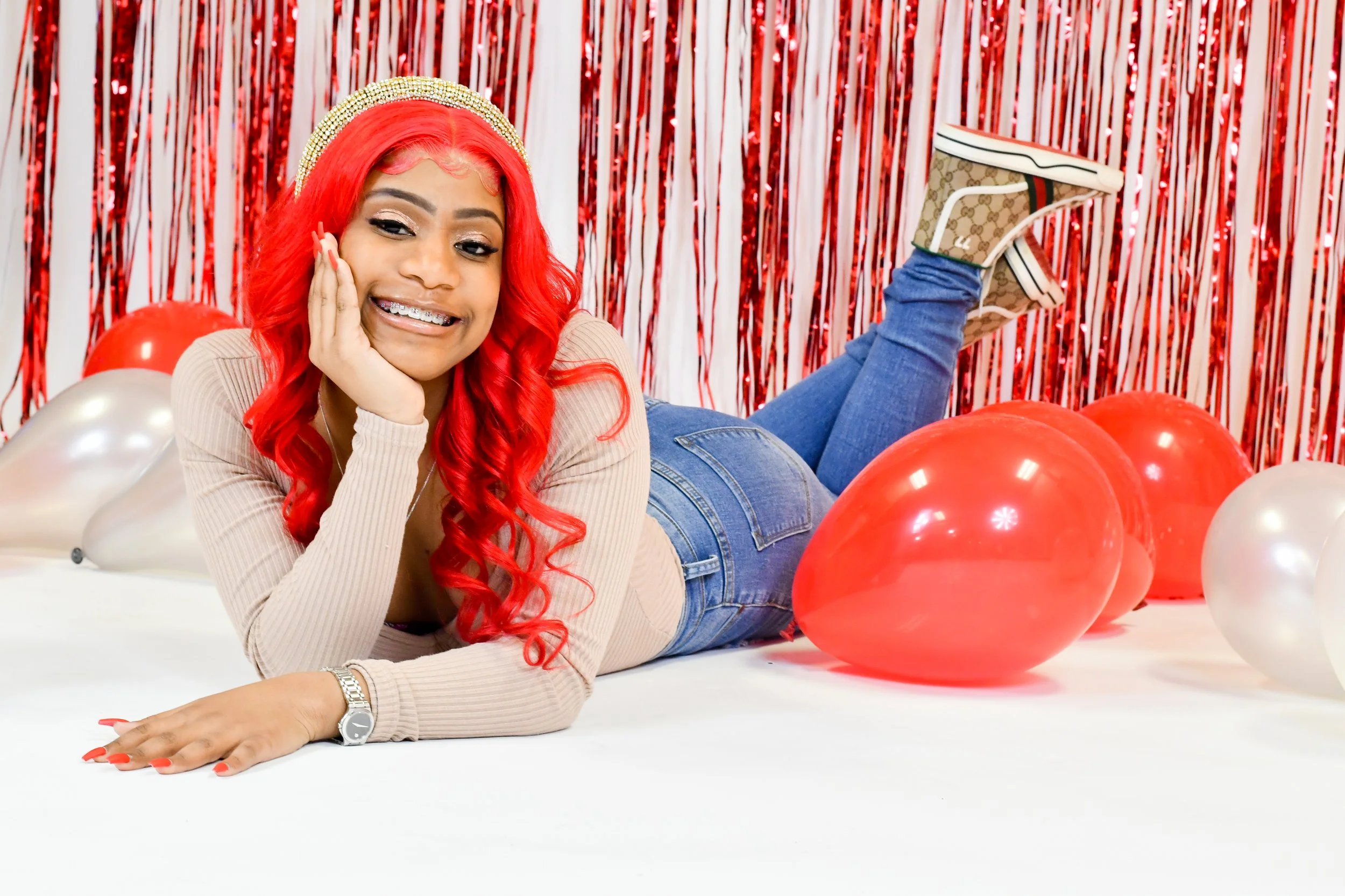 Person with red hair and braces lying on the floor surrounded by red and silver balloons, with a red metallic fringe background.