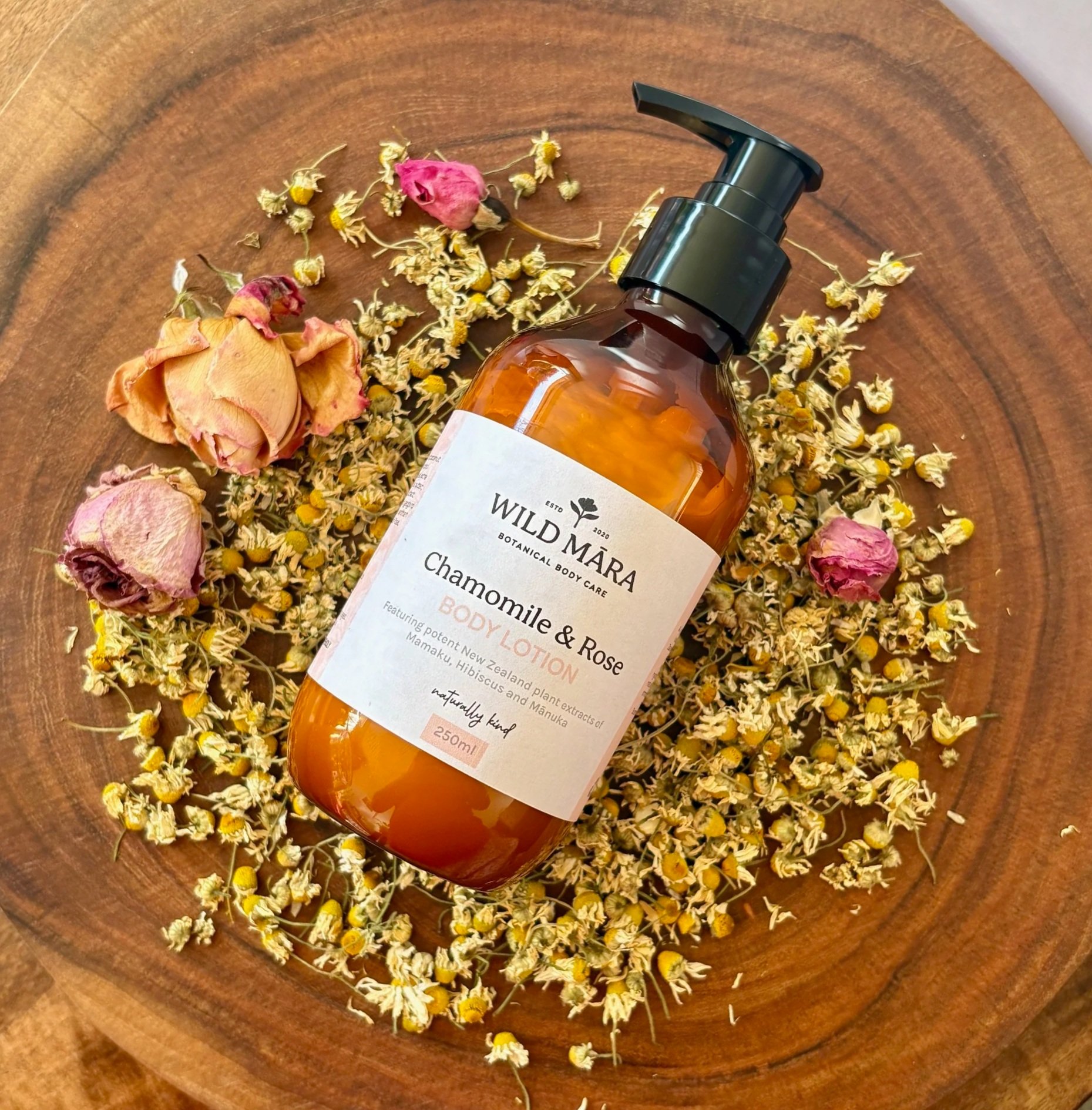 A bottle of botanical body care lotion labeled 'Wild Māra' with chamomile and rose scent, lying on a wooden surface surrounded by dried chamomile flowers and pink rosebuds.