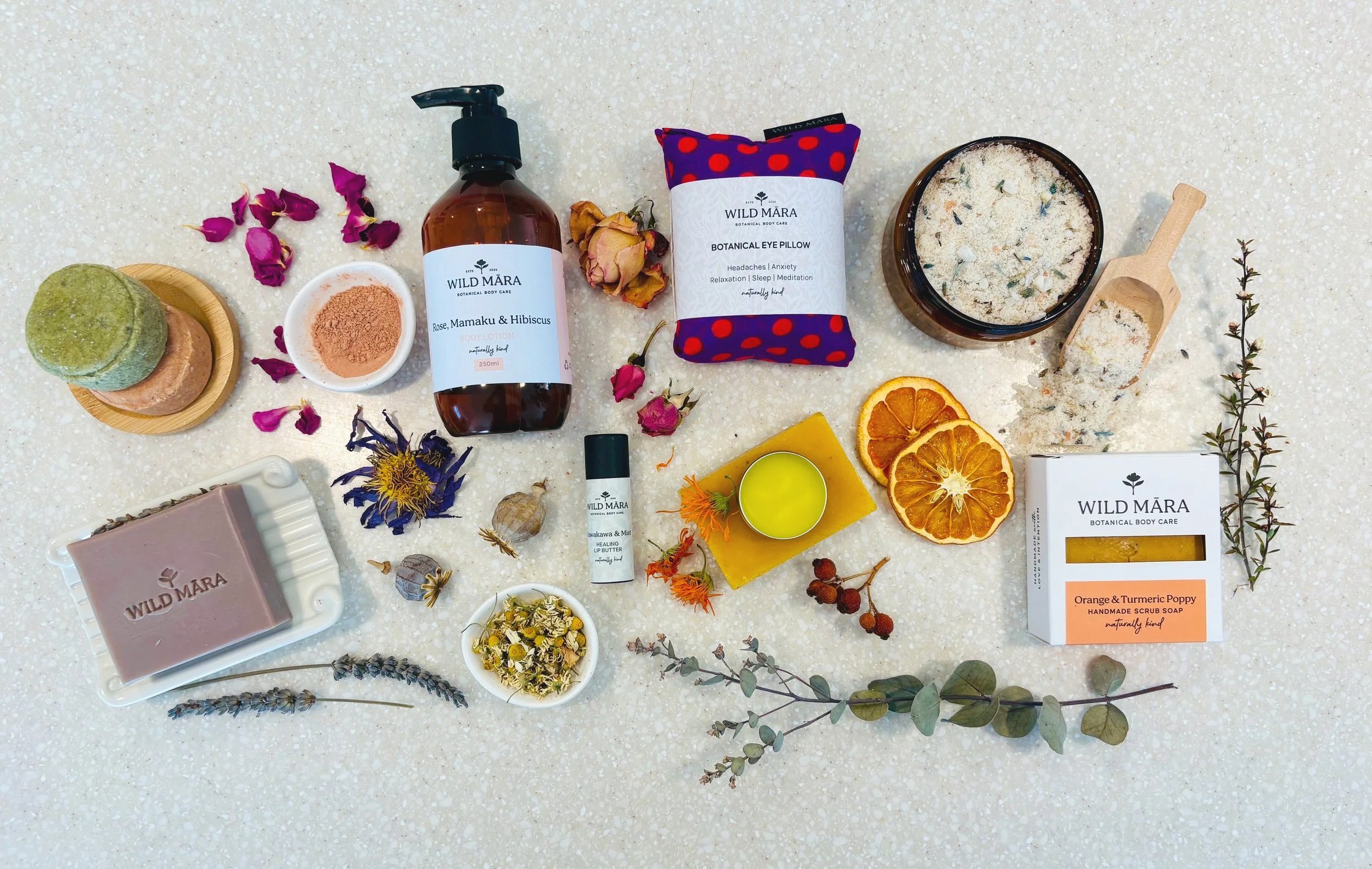 An assortment of natural and organic skincare and body care products, including soap, body lotion, face pillow, bath salts, dried flowers, dried citrus slices, and herbal sprigs, arranged on a white speckled surface.