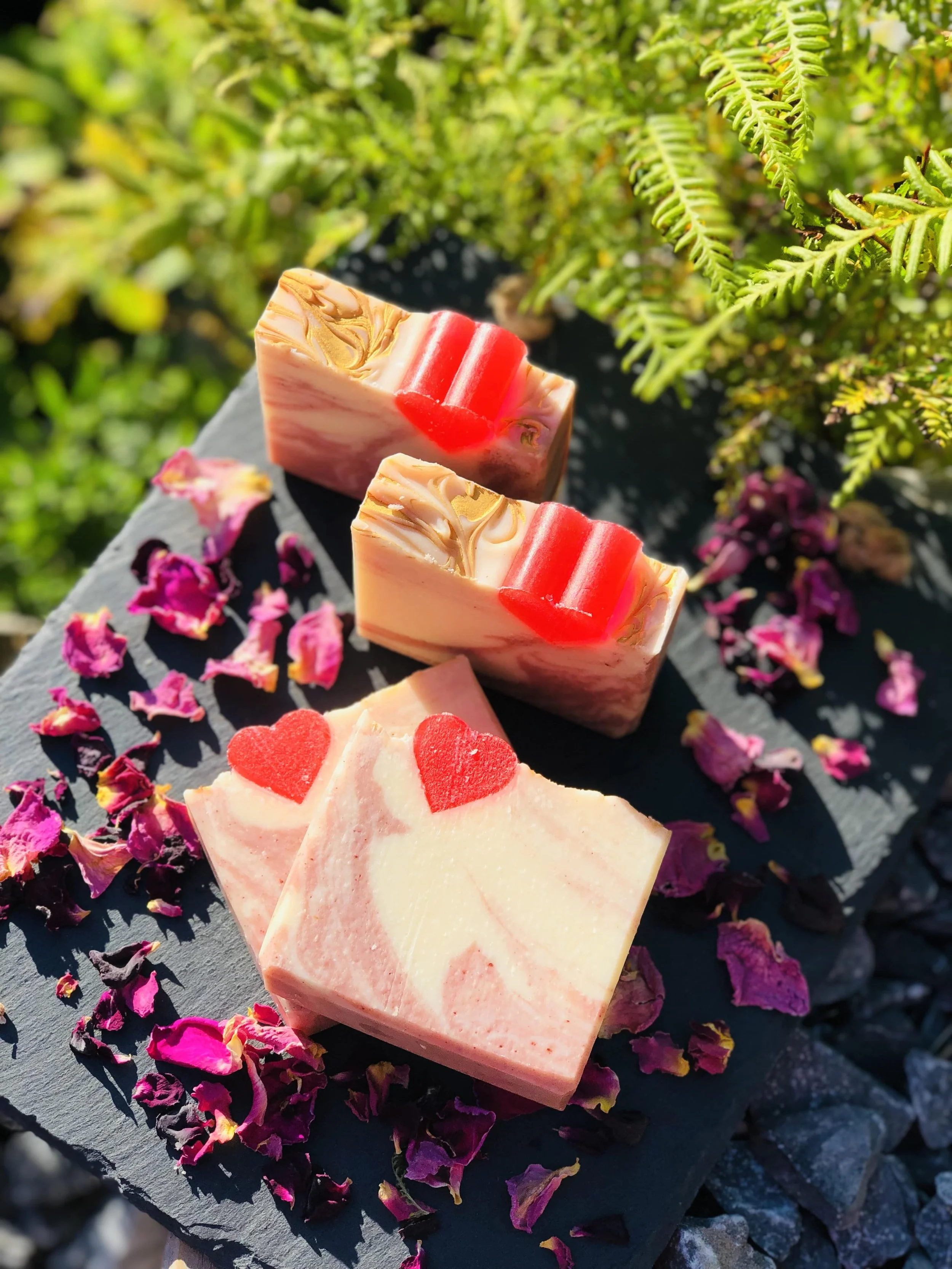 Three handcrafted soap bars on black slate with pink flower petals and green ferns in the background.