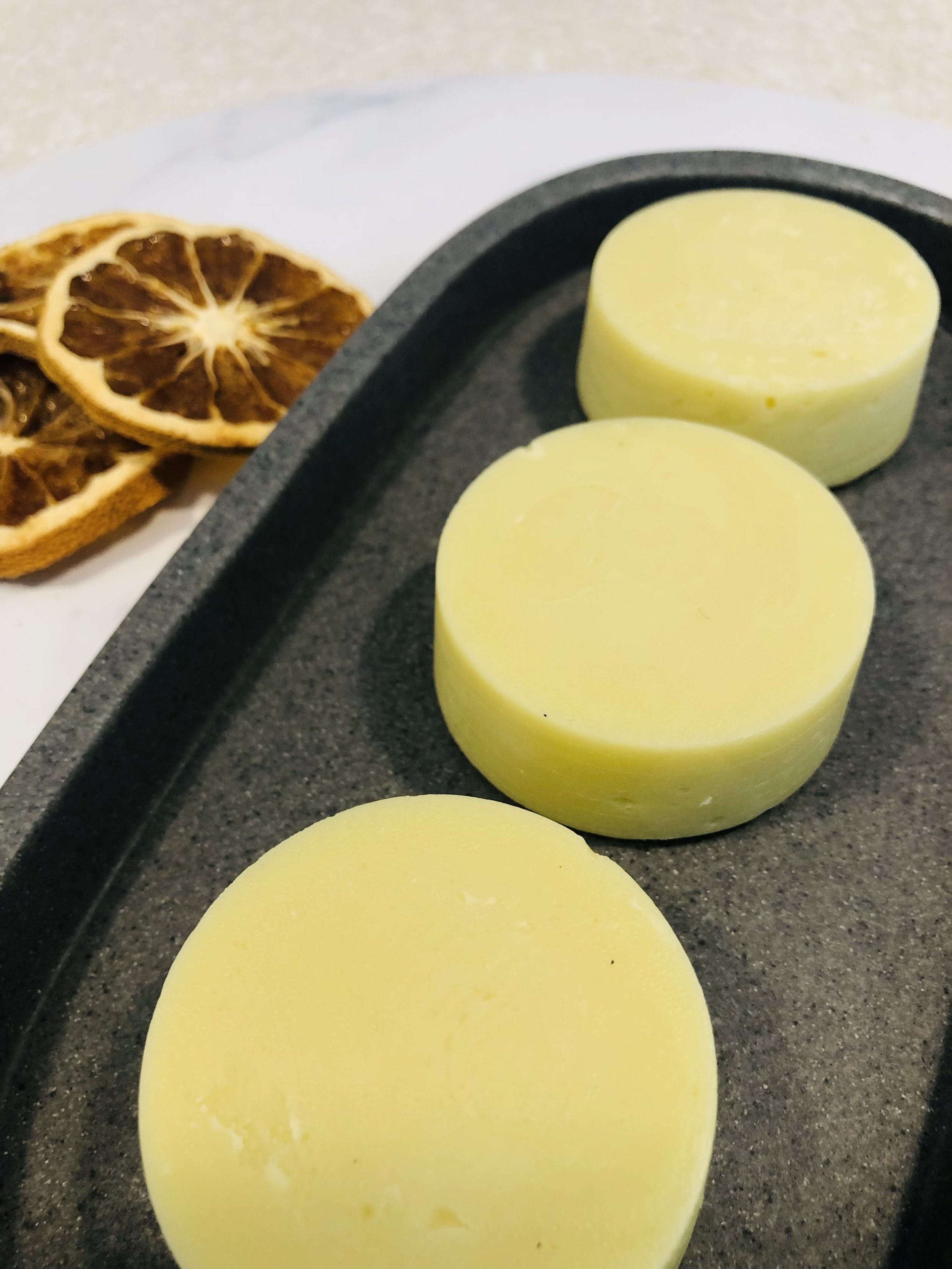 Round blocks of yellow conditioner bars on a black soap dish, with dried orange slices on a white background nearby.