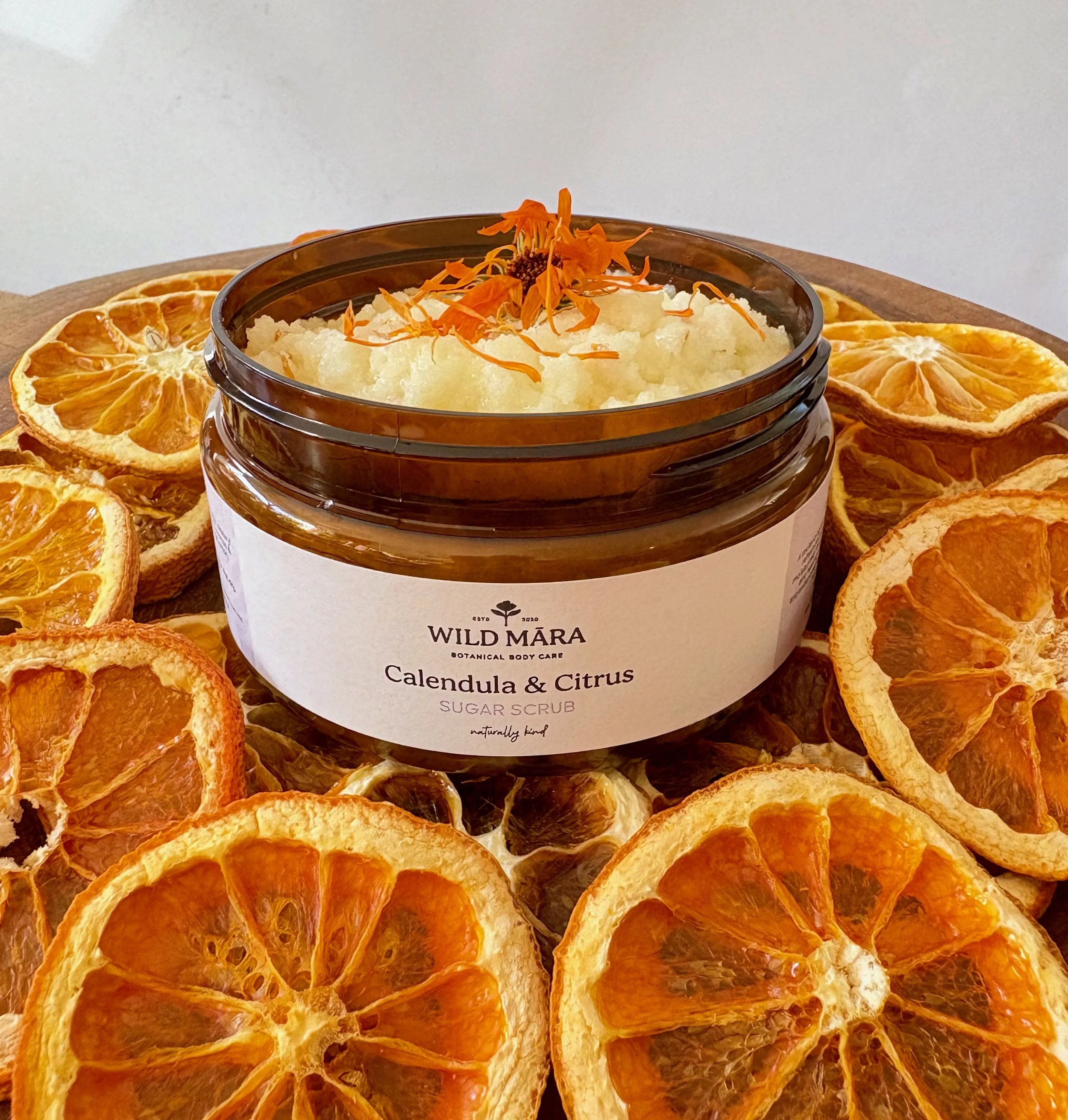 A container of Wild Mara Calendula & Citrus sugar scrub surrounded by dried orange slices.