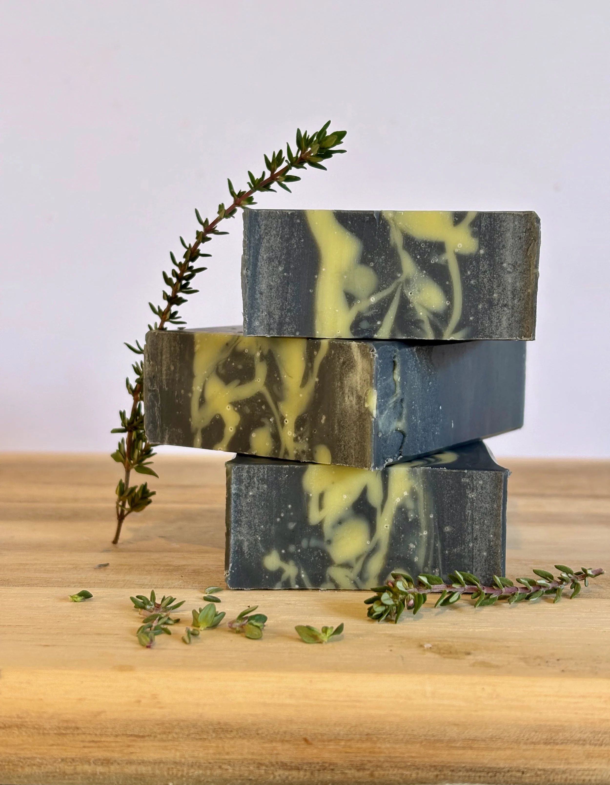 Stacked blocks of soap with a sprig of green herb on top, placed on a wooden surface with loose herb pieces around.