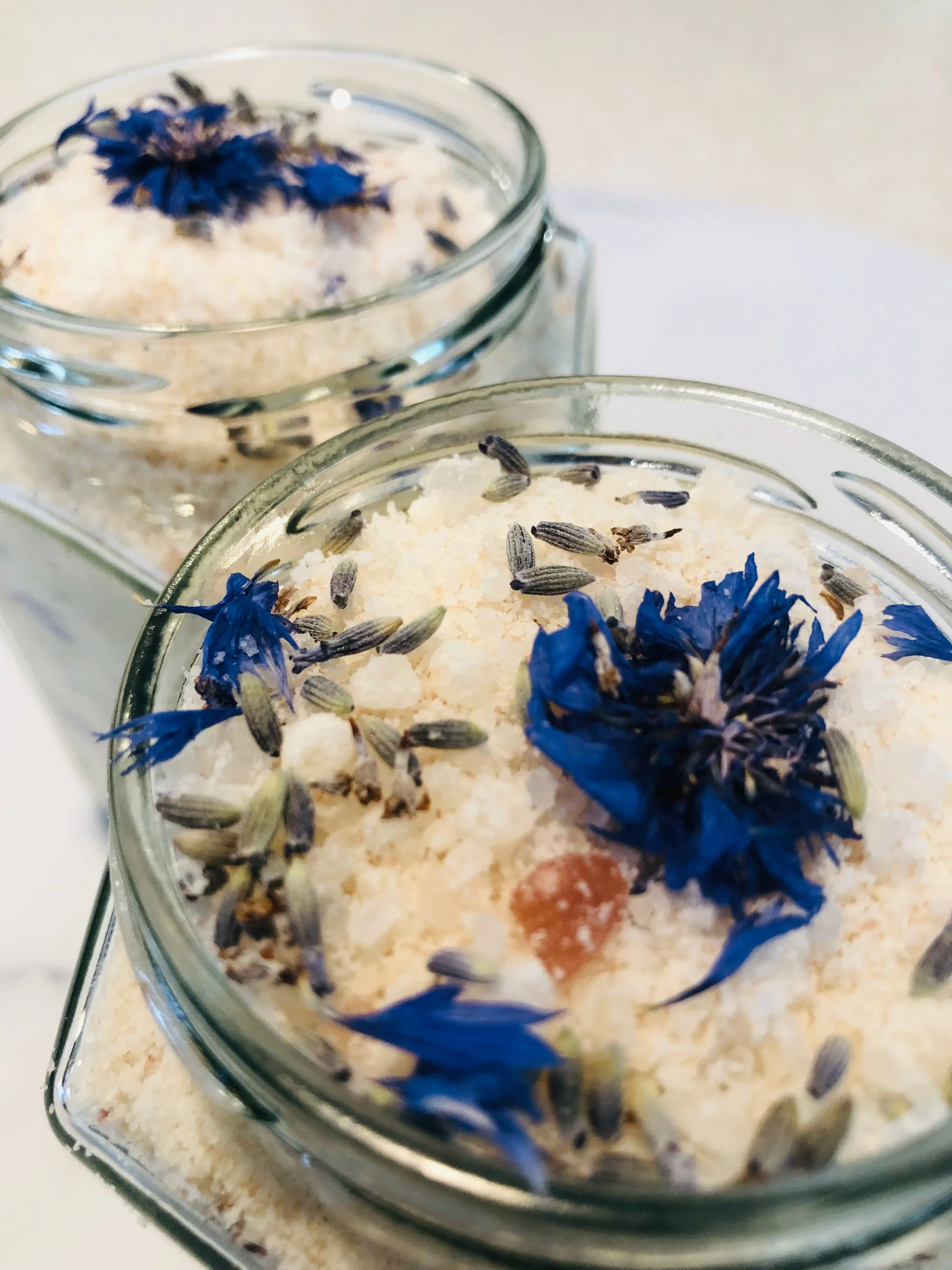 Close-up of two glass jars filled with a white, flaky substance, decorated with dried blue flowers and lavender buds.