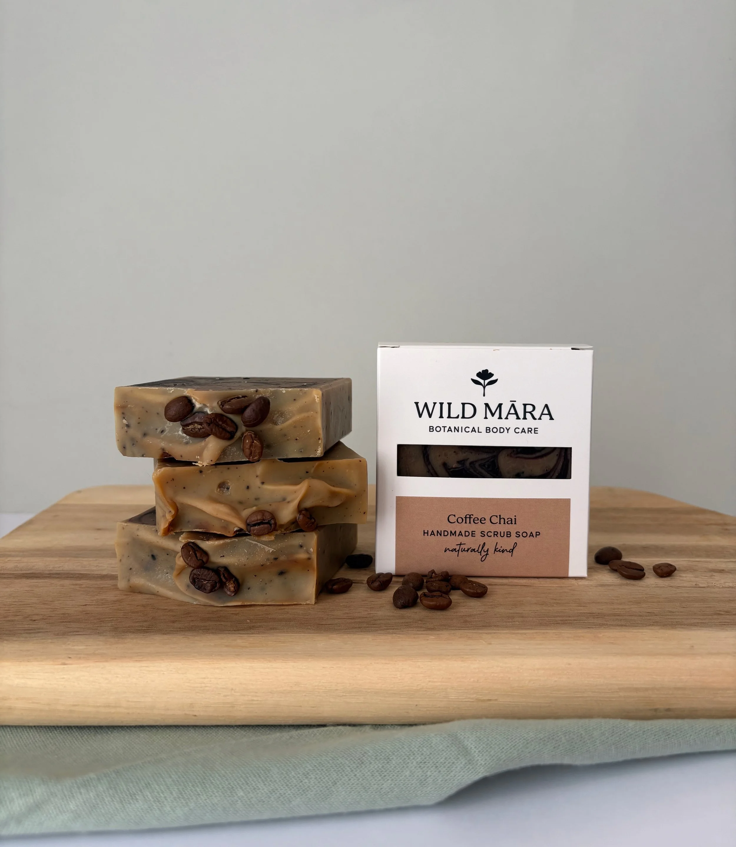 Stacked handmade soap bars with coffee beans, next to a white box labeled WILD MÄRA Botanical Body Care Coffee Chai Handmade Scrub Soap on a wooden surface.