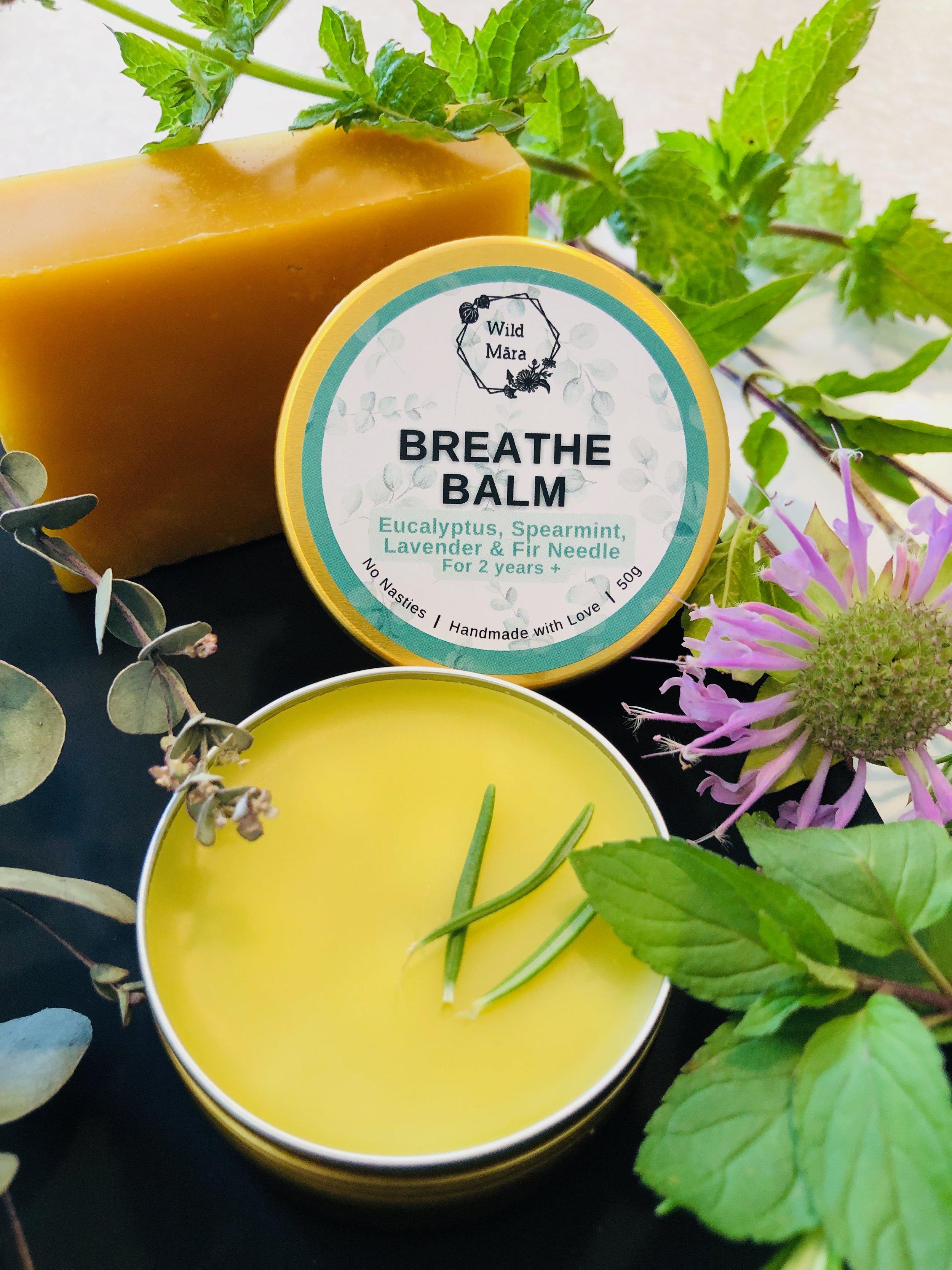 A round yellow tin labeled "Breathe Balm" with eucalyptus, spearmint, lavender, and fir needle ingredients, accompanied by a bar of soap, greenery, and a purple flower on a black surface.