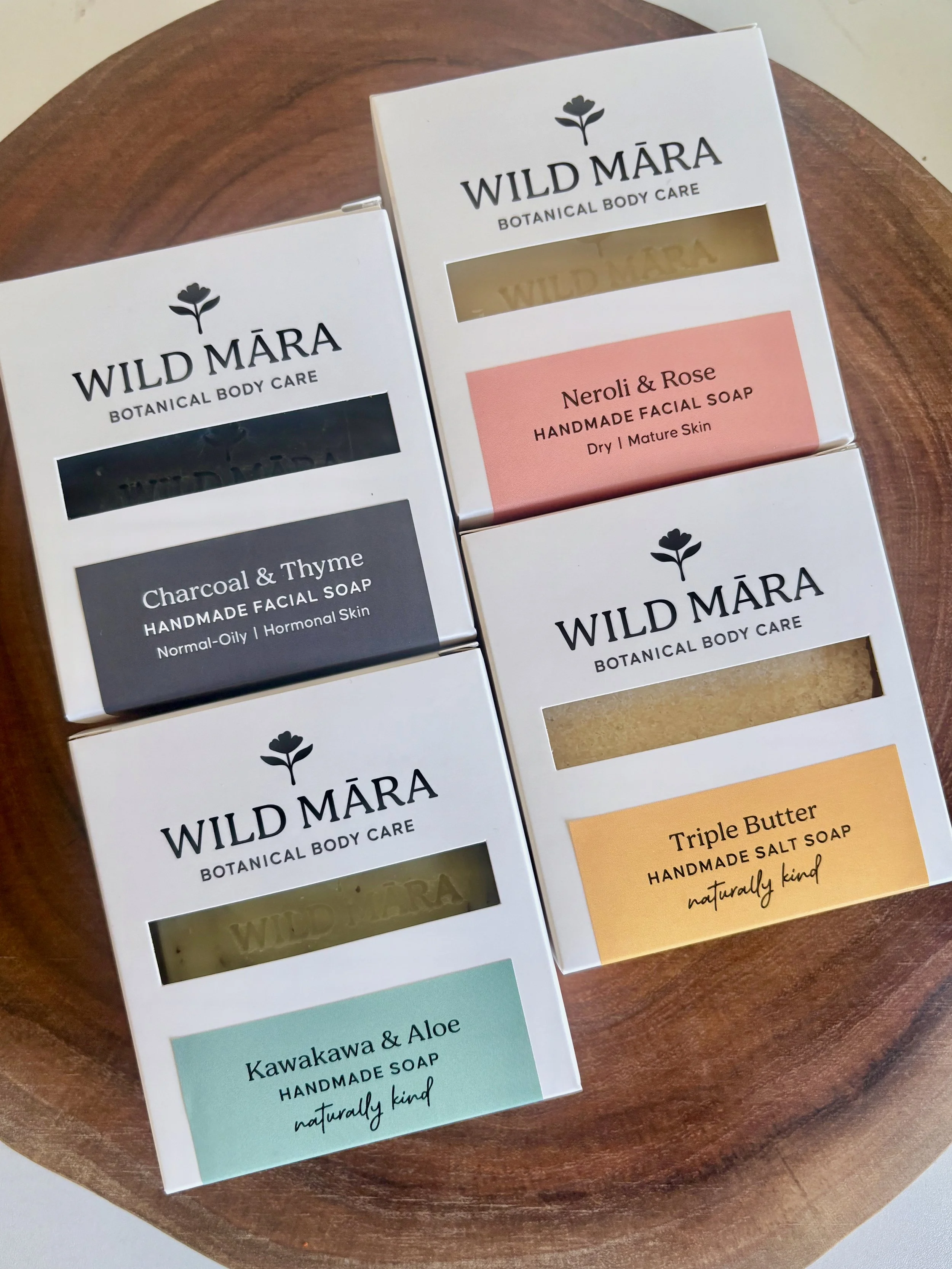 Several boxes of Wild Mára Botanical Body Care handmade soaps on a wooden surface. The flavors include Charcoal & Thyme, Neroli & Rose, Kawakawa & Aloe, and Triple Butter, with descriptions for each.