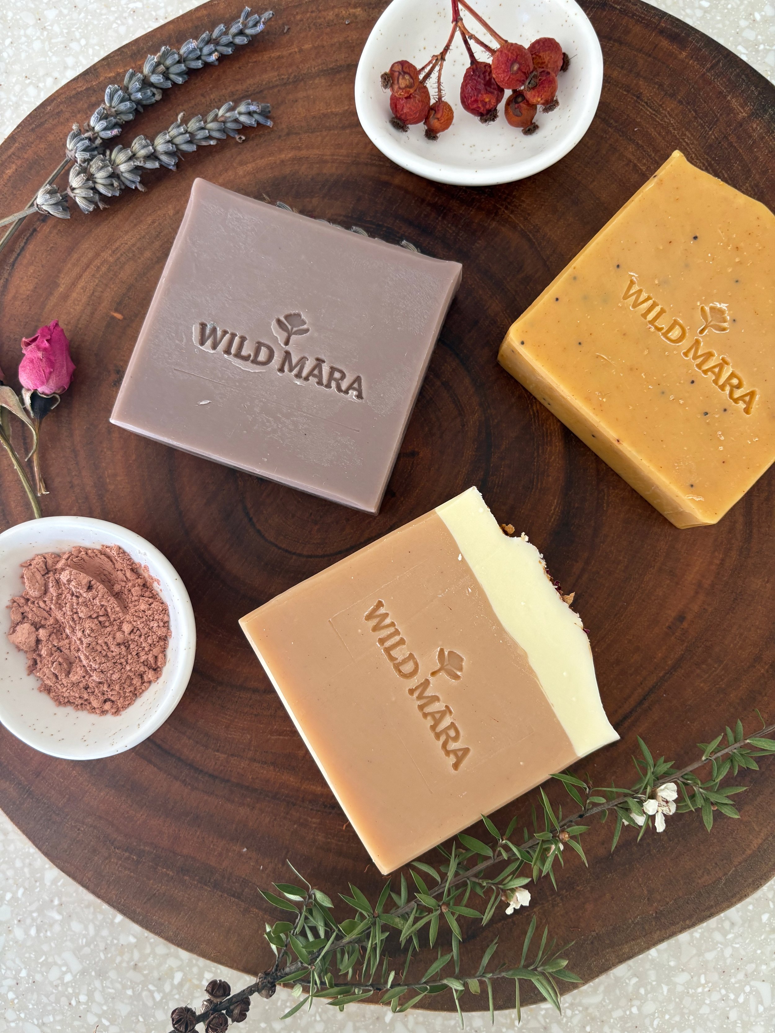 Three artisanal soap bars labeled 'WILD MĀRA' in different colours, a small dish with dried rosehips, a sprig of lavender, a small bowl of clay, and a sprig of greenery on a wooden tray.