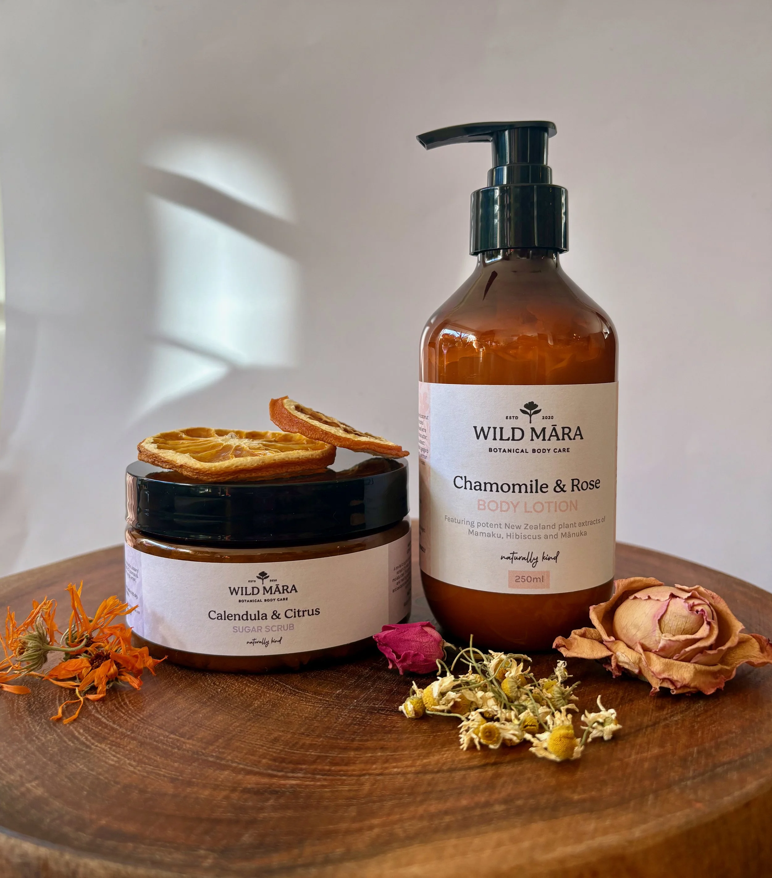 A brown bottle of Wild Mara botanical body care chamomile and rose body lotion surrounded by dried herbs, flowers, and slices of dried citrus on a wooden surface.