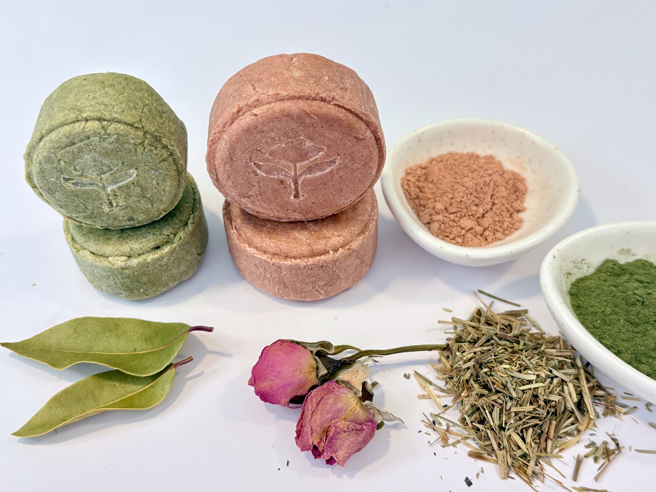 A collection of natural products including three green matcha stone tablets, three pink Hakka mudstone tablets with a scorpion imprint, dried rosebuds, dried green leaves, ground herbs in small white dishes, and dried herbs and twigs on a white background.