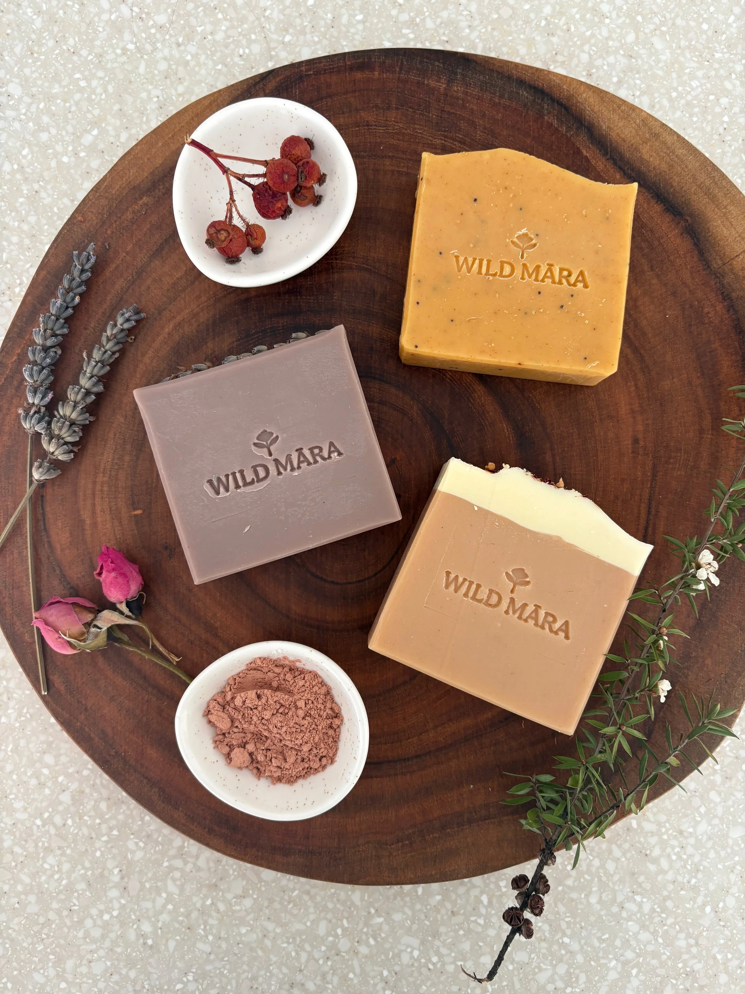 Three artisanal soap bars labeled 'WILD MARA' in different colors, a small dish with dried berries, a sprig of lavender, a small bowl of cocoa powder, and a sprig of greenery on a wooden tray.