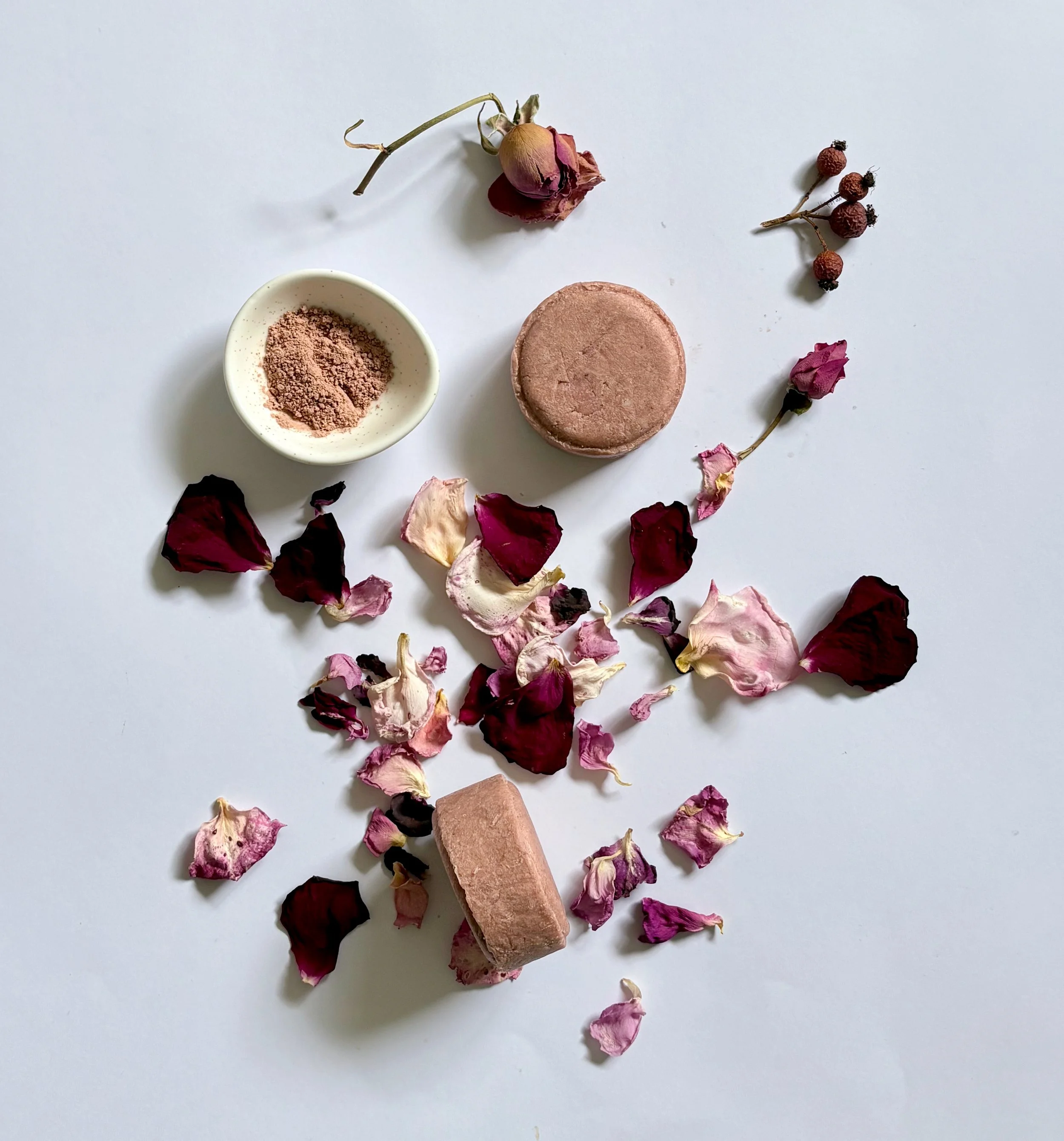 A white surface with dried rose petals, a pink rose, a round pink bath bomb, a small white bowl with pink powder, and several dried berries.