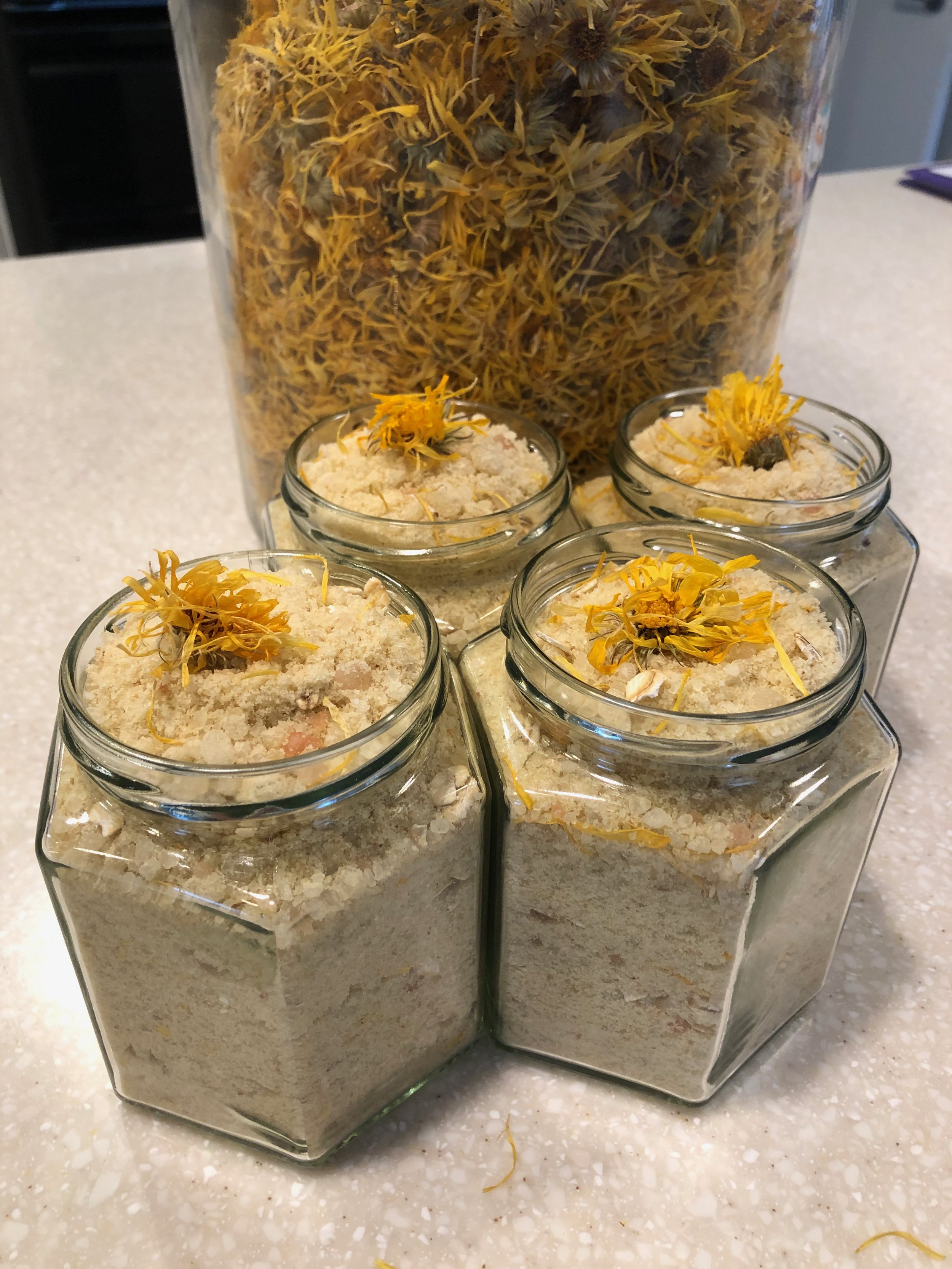 Four glass jars filled with a white, crumbly substance, each topped with yellow dried flowers, in front of a large clear container filled with dried yellow and brown flowers.