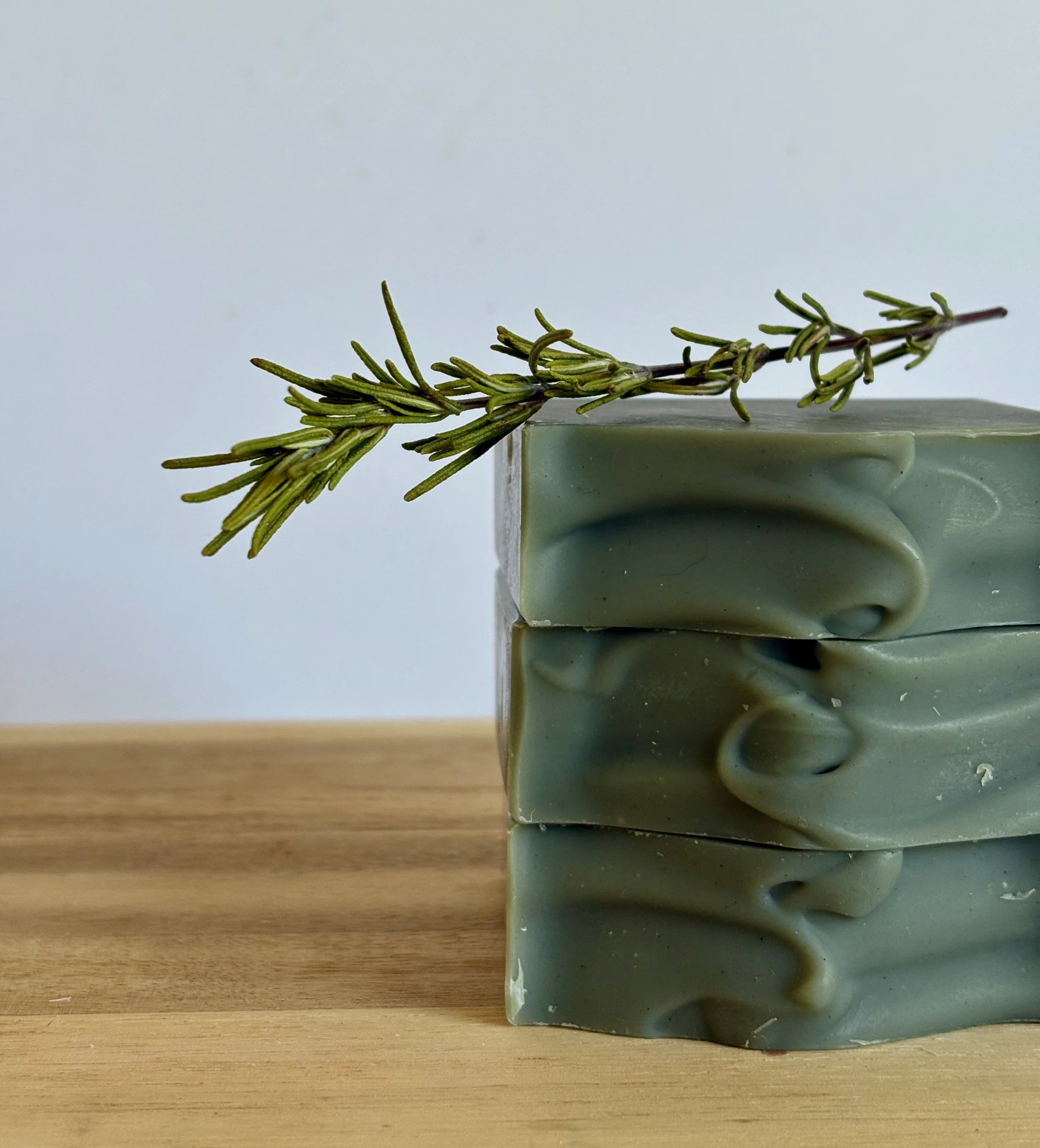 Close-up of a small, irregularly shaped block of greenish-grey soap with a wavy textured surface, placed on a wooden surface. A sprig of fresh rosemary rests atop the soap.