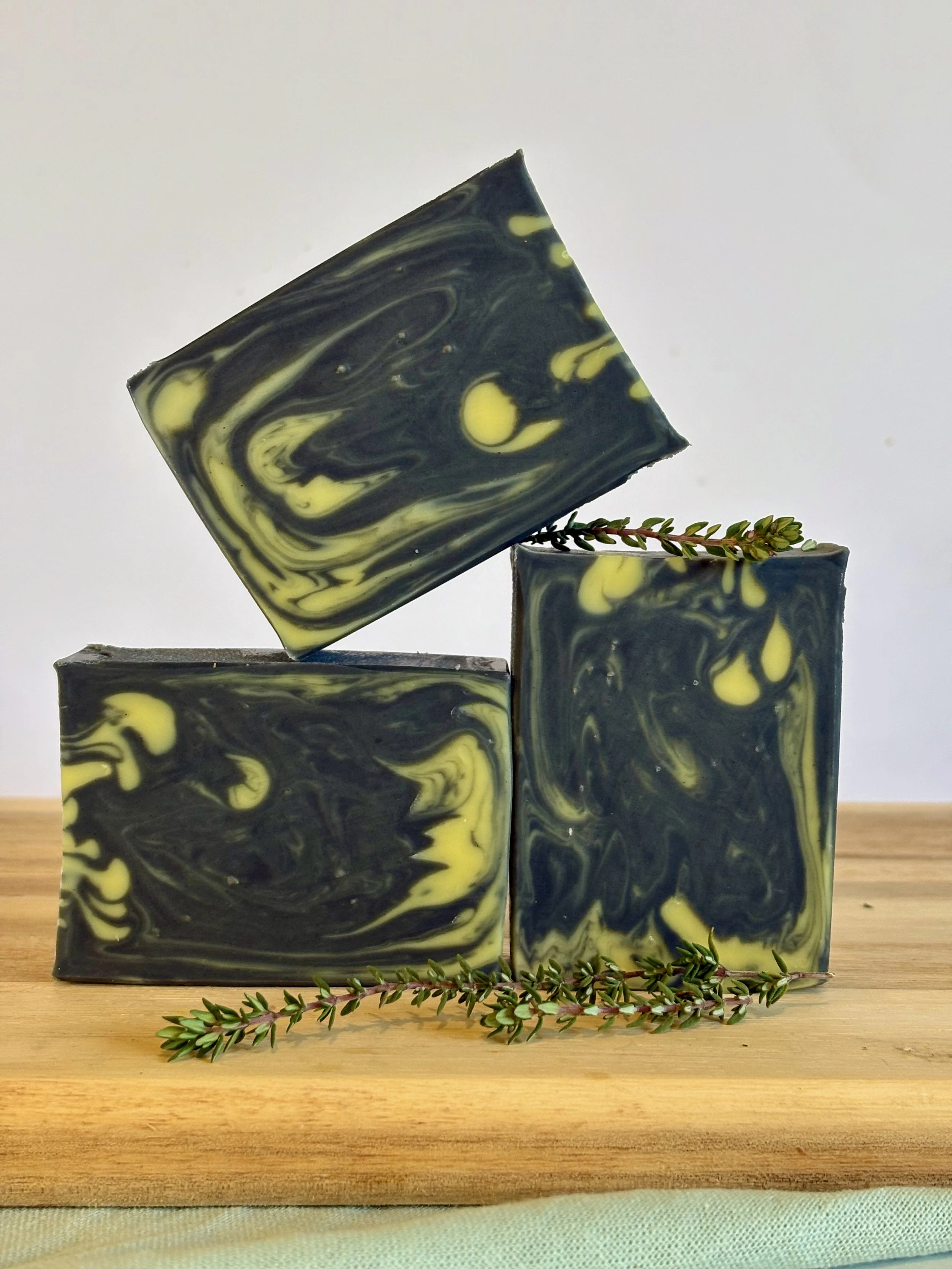 Three marbled soap bars with yellow and black swirls, placed on a wooden surface with sprigs of greenery.