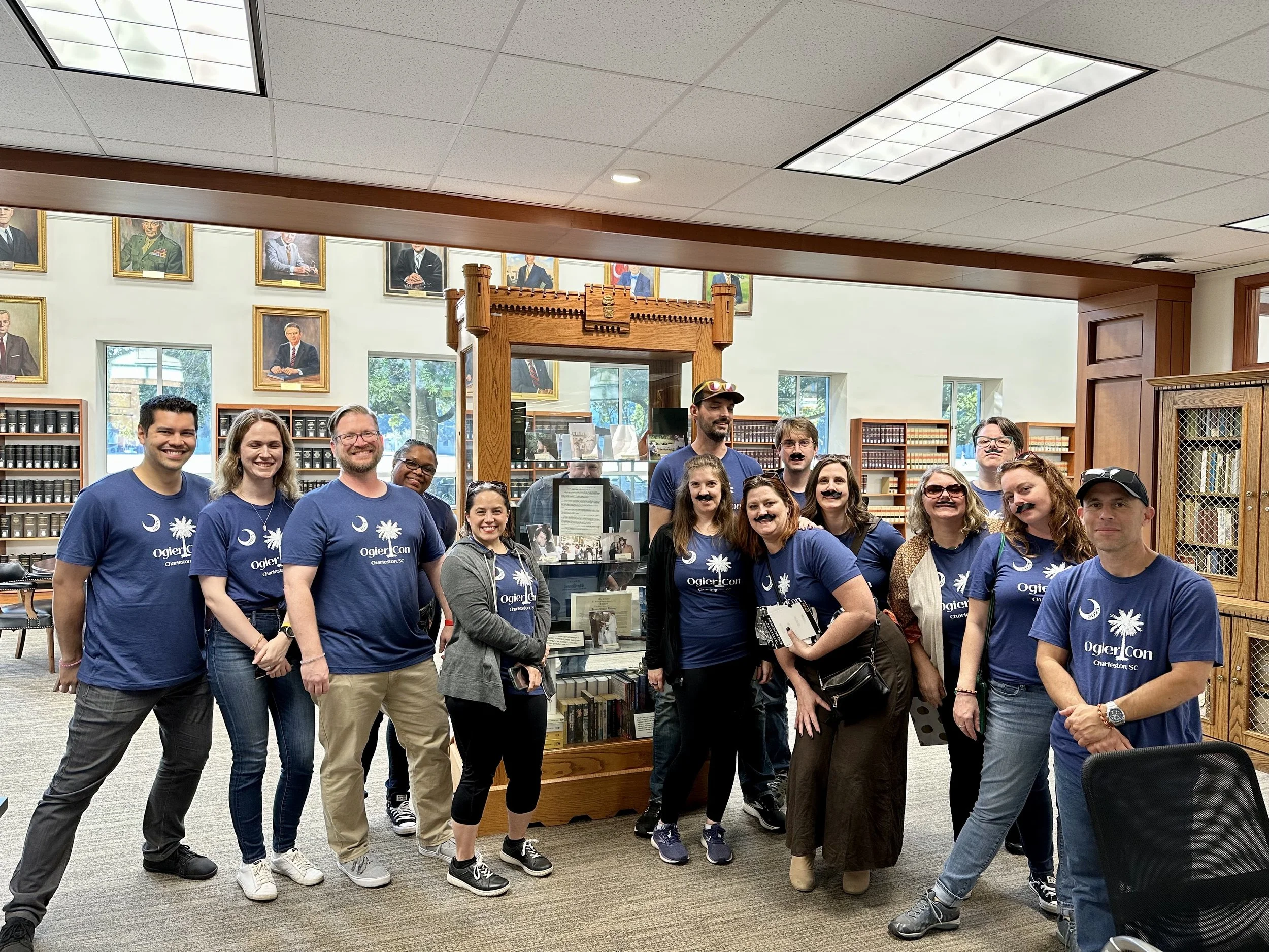 A group of fans wearing blue OgierCon tees stand in a row in brightly lit library. At the center is a tall glass display case with crenelated wooden trim that contains memorabilia related to Robert Jordan and the Wheel of Time.
