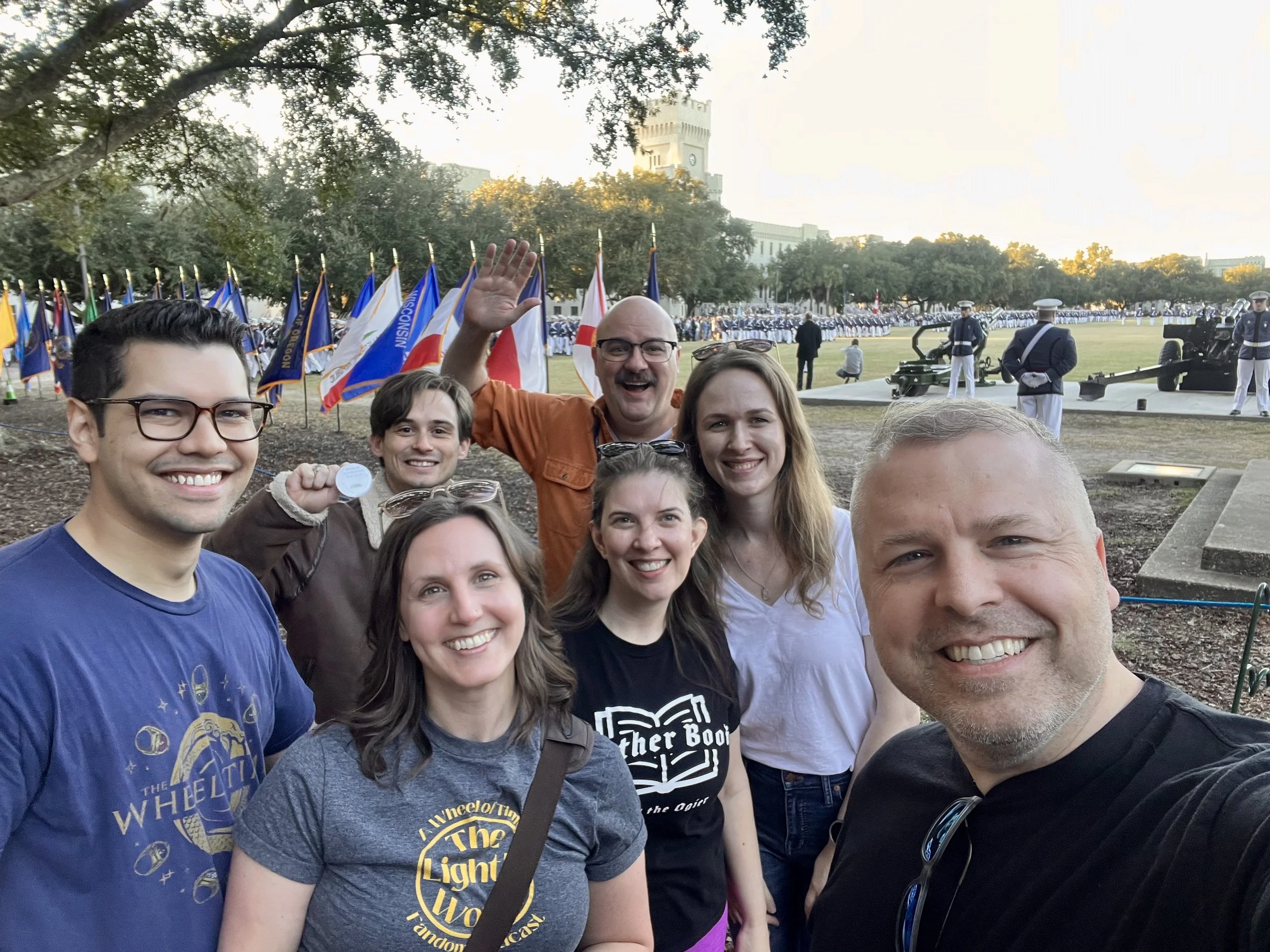 A selfie of 7 people in front of several colorful flags outlining a large green field. To the right of the flats are two cannons and cadets in uniform. At the back of the selfie is Michael Livingston smiling and waving.