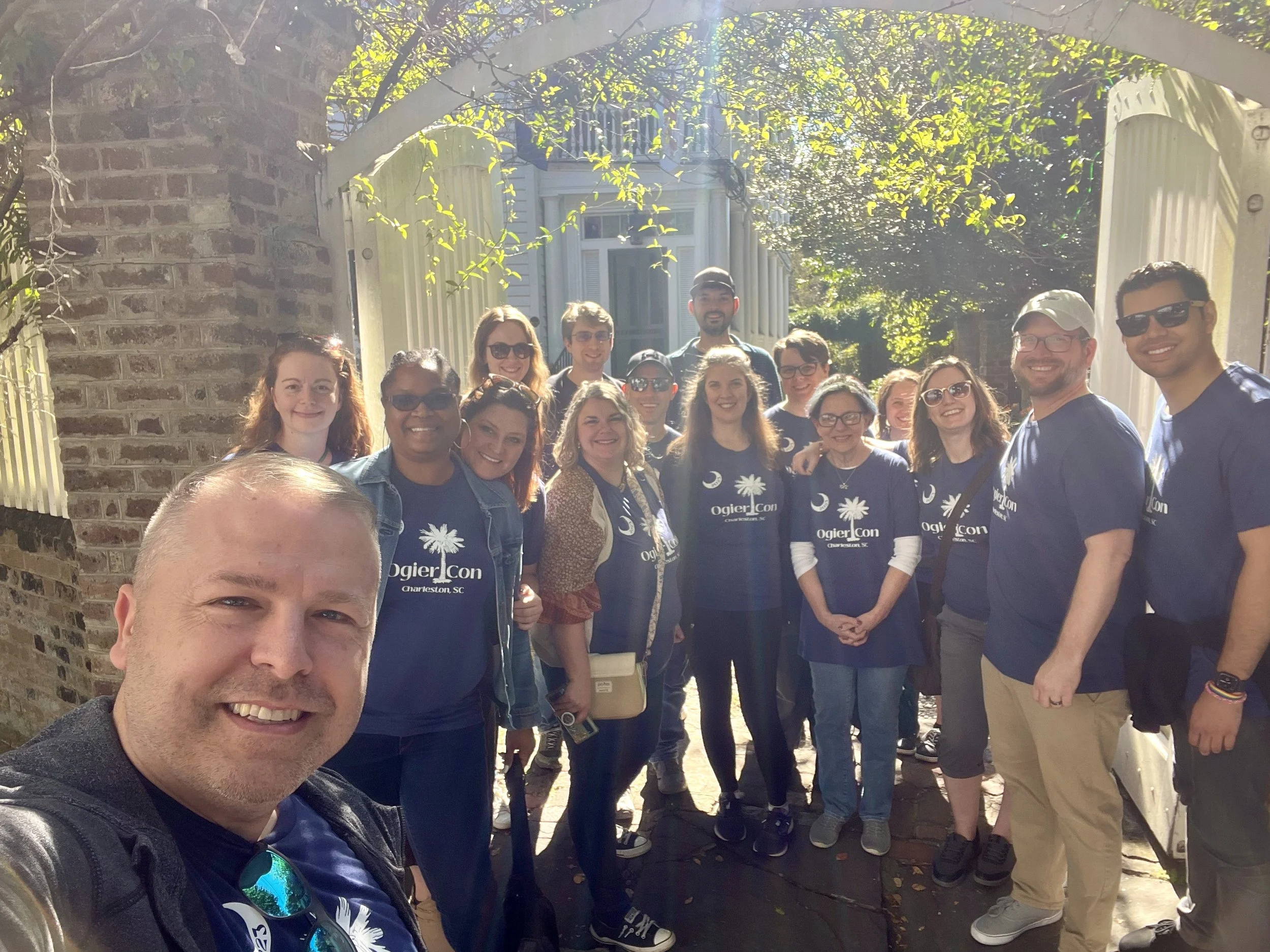 Brightly sunlit selfie of a group of fans outside the white gates to Robert Jordan's estate. The group all wear blue OgierCon tees, including Maria Simons who is front and center, wearing a tiara.