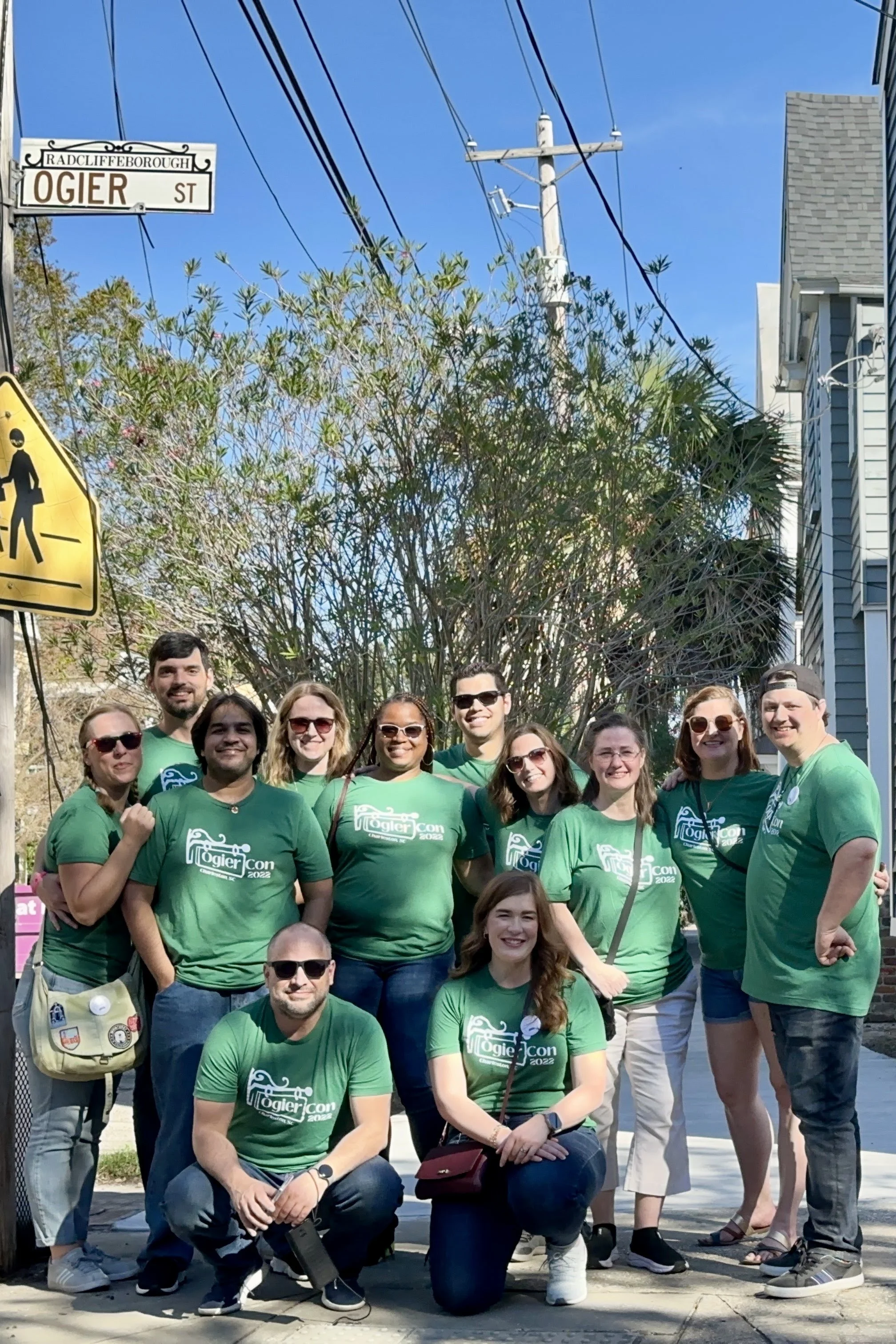 A group of 12 people wearing green OgierCon tees stand crowded together on the sidewalk on Ogier Street in Charleston. Above the group's head on a phone pole is a white street sign with "Ogier St." in black lettering