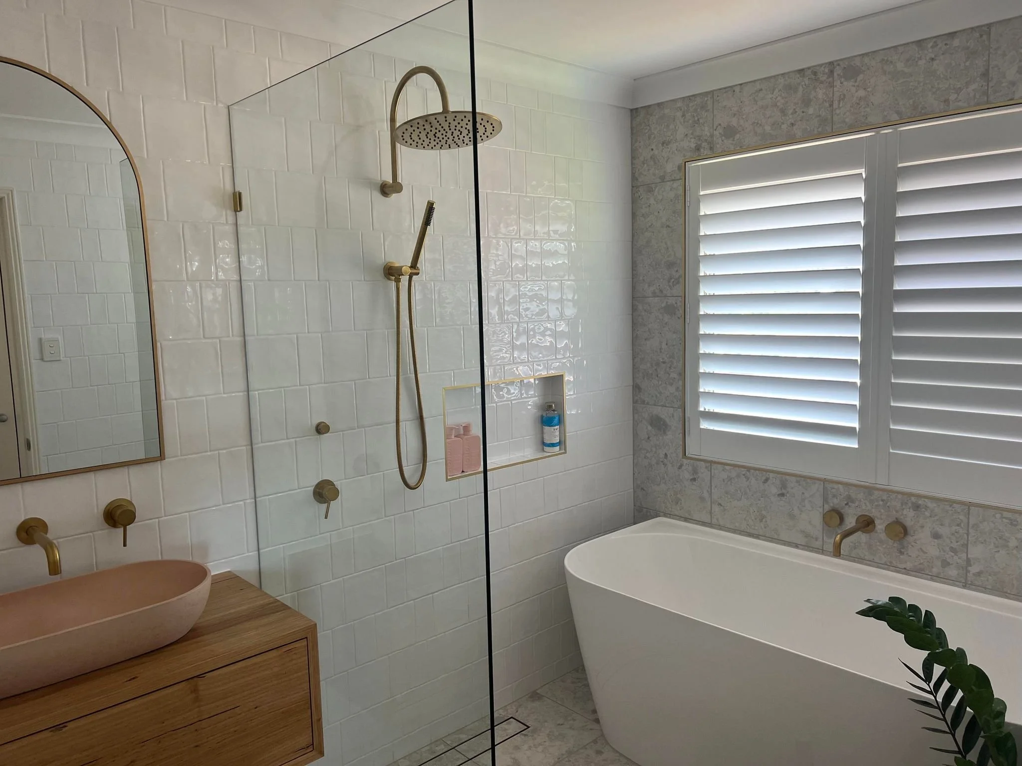 Modern bathroom with a brass overhead shower, a handheld shower, white tile walls, a window with white shutters, a bathtub, a wooden vanity with a stone basin, and some green plants.