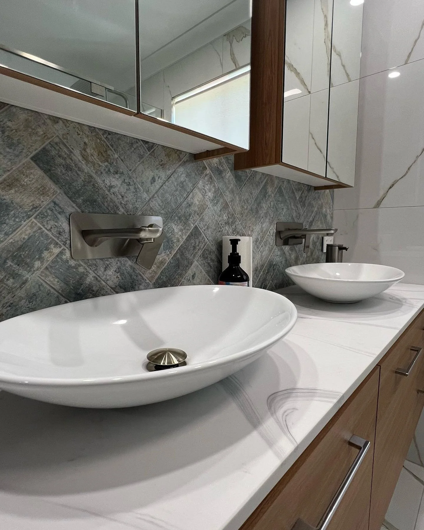 A modern bathroom vanity with two white vessel sinks, a single handle faucet, a soap dispenser, and a mirror cabinet. The countertop is white with a wood-finish cabinet below. The background features a tiled wall with marble pattern and another tiled wall with a window.