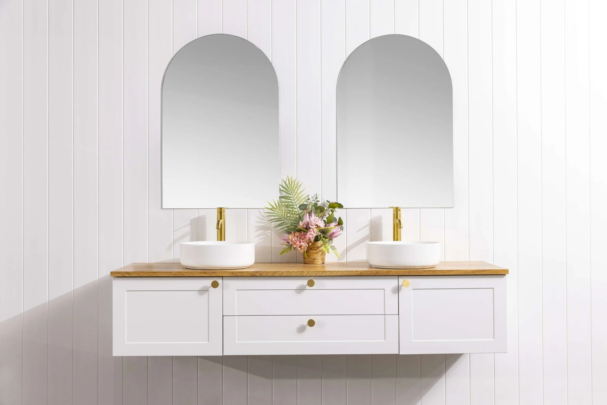 Bathroom vanity with two vessel sinks, gold faucets, a wooden countertop, two mirrors, and a flower arrangement in a basket against a white paneled wall.