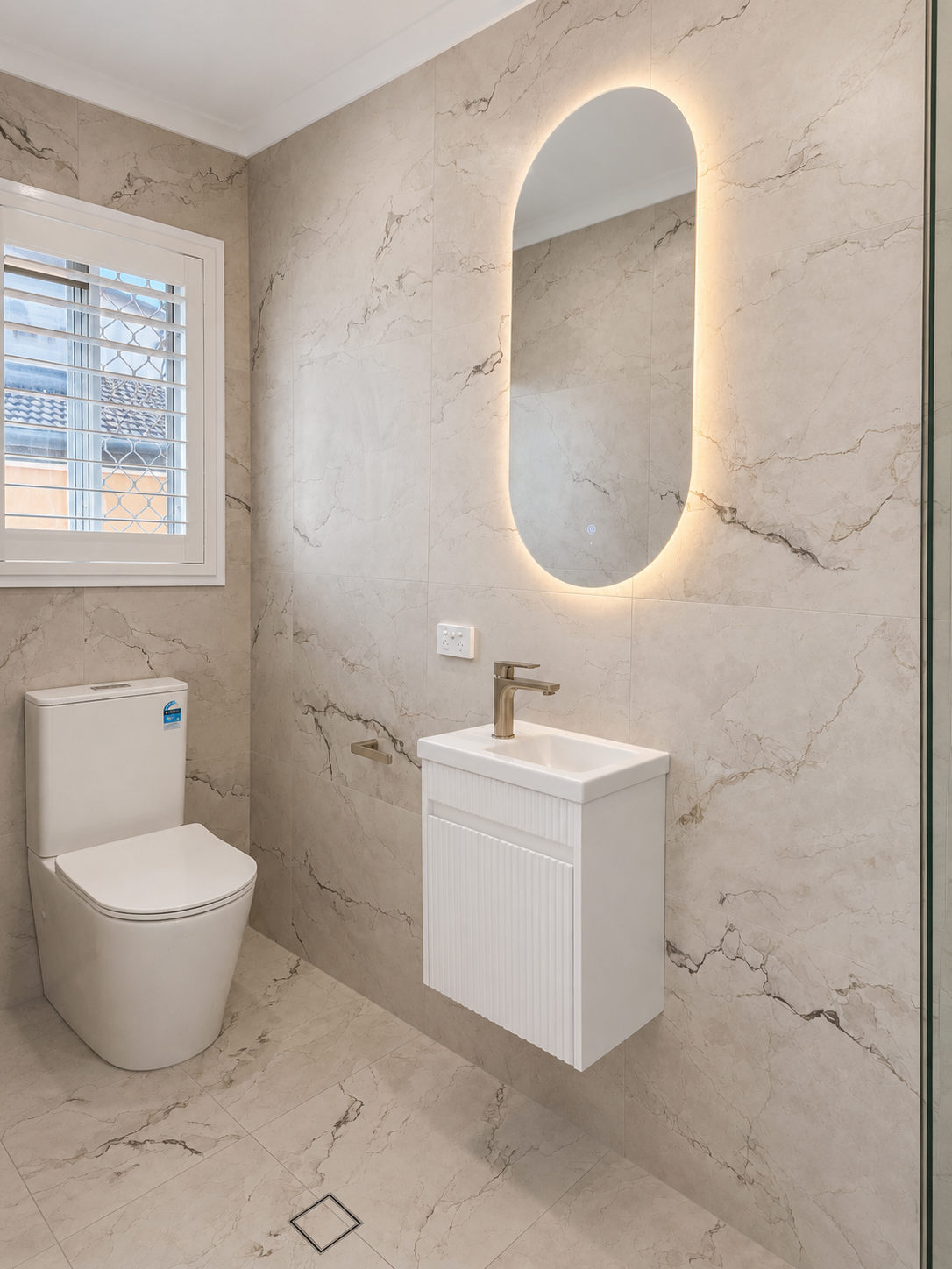 Modern bathroom with beige marble walls and floor, a white toilet, small white sink with brass faucet, illuminated oval mirror, window with white blinds, and a square floor drain.