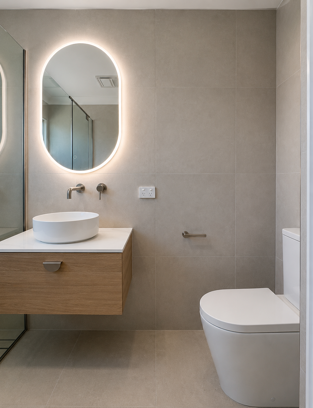 Modern bathroom with beige tiled walls, a backlit oval mirror, a white vessel sink on a wooden vanity, wall-mounted faucet, and a toilet.