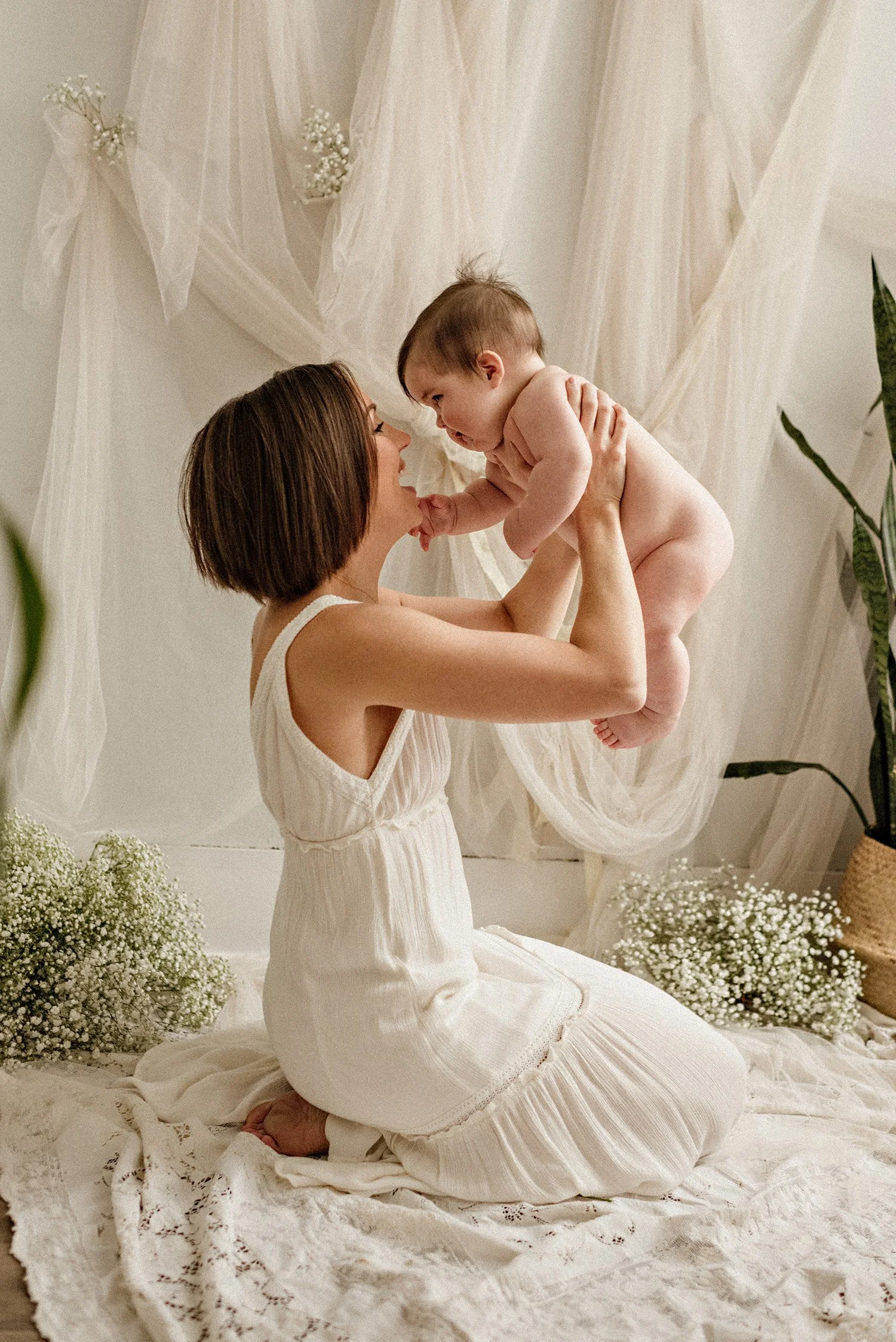 A woman in a white dress kneeling on a floor decorated with white fabric and baby's breath flowers, holding a baby. The background features sheer white drapes and plants.