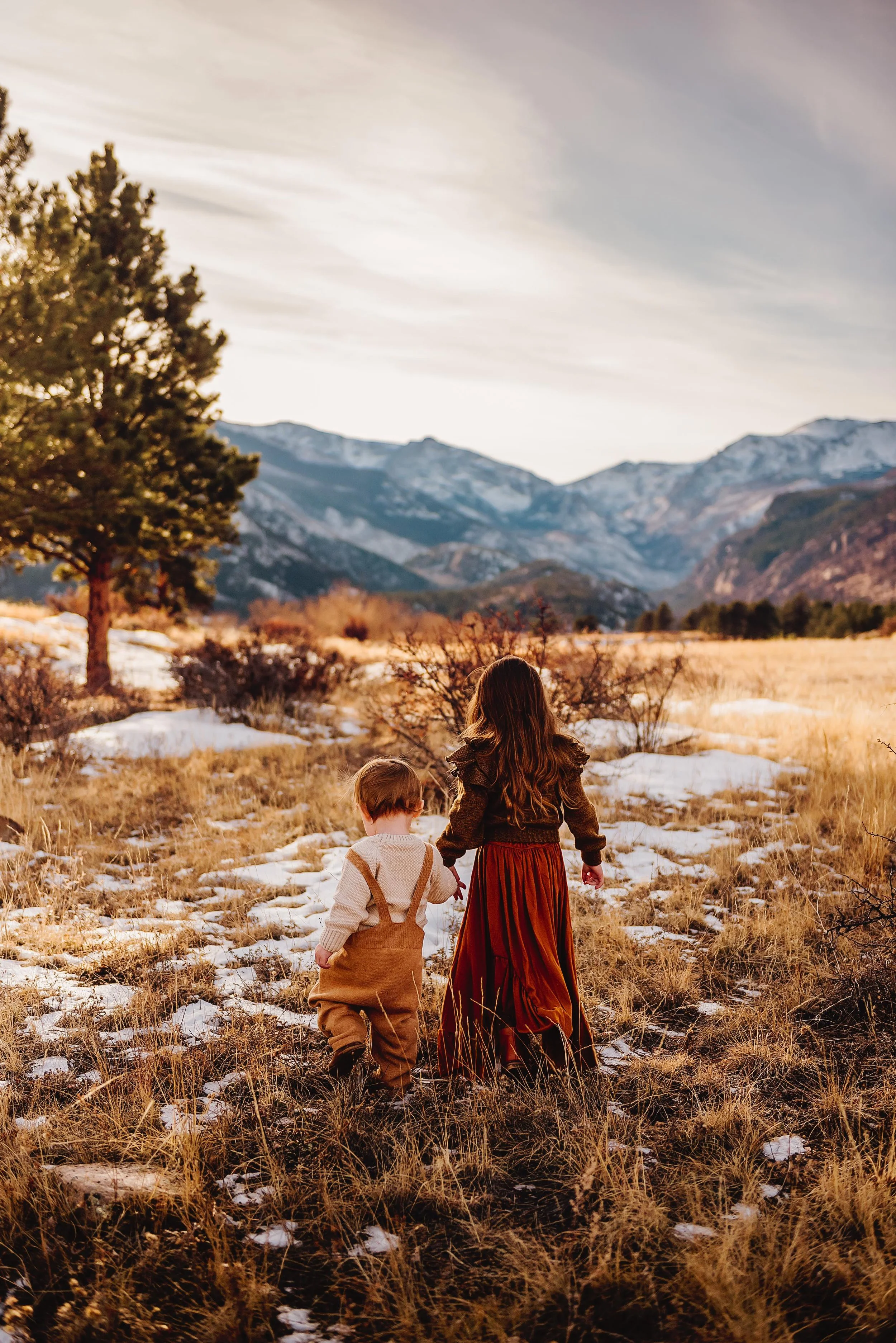 Two children walking hand-in-hand through a snowy field with mountains in the background during sunset.