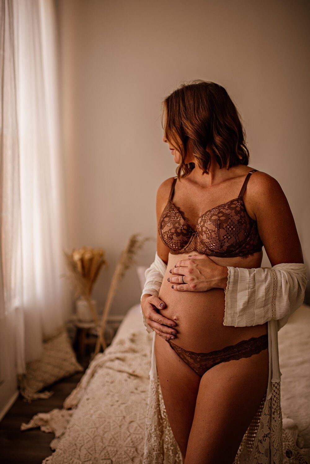 Pregnant woman in brown lingerie holding her belly, standing in a softly lit bedroom with neutral decor.