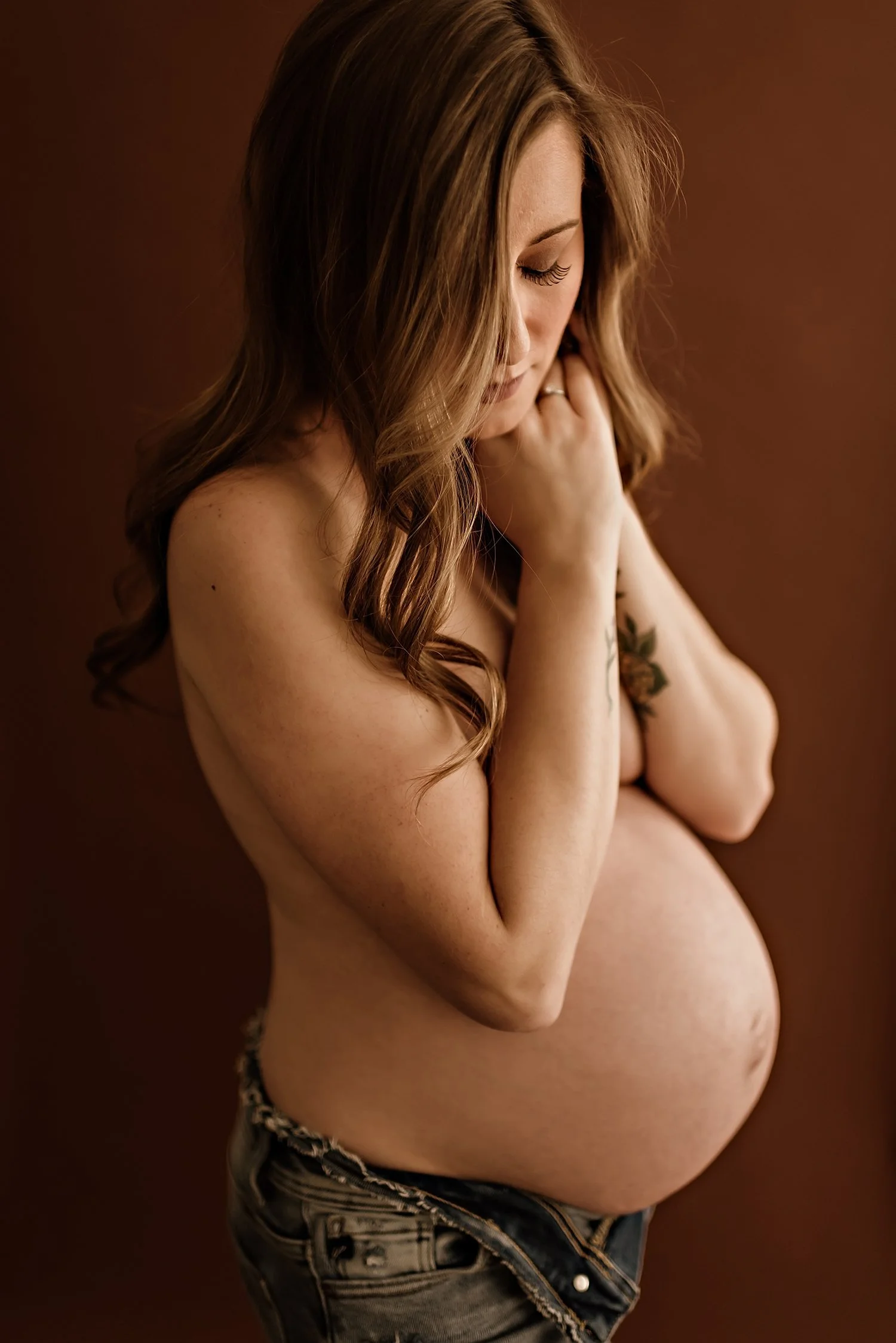 Pregnant woman posing in jeans against a brown background.