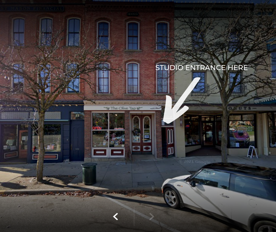 Street view of a brick building with "The Olive Tap" store and adjacent shops. An arrow indicates a studio entrance next to the store. A white car is parked on the street.