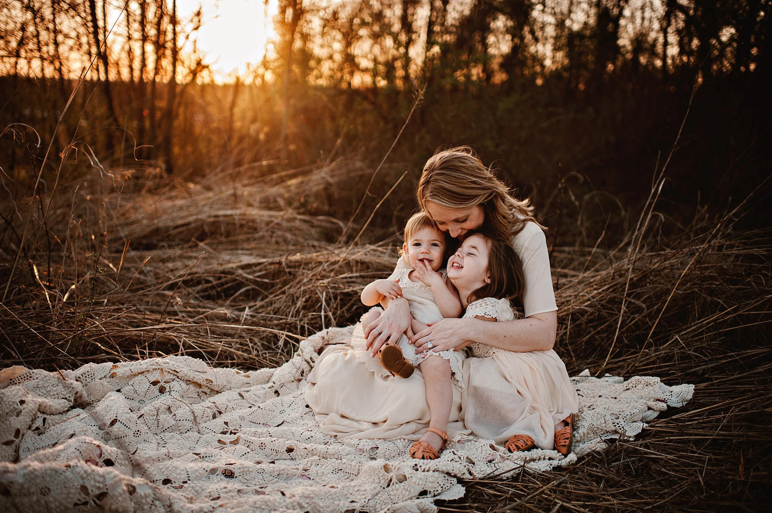 A mother embraces two children sitting on a crochet blanket in a field during golden hour.