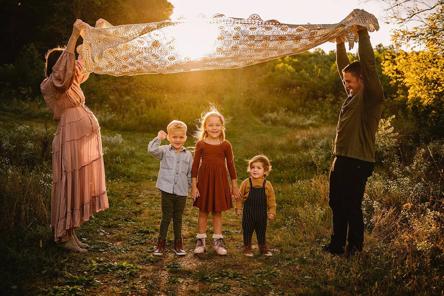 Family with three children standing under a lace cloth, held by two adults, in a sunny outdoor setting.