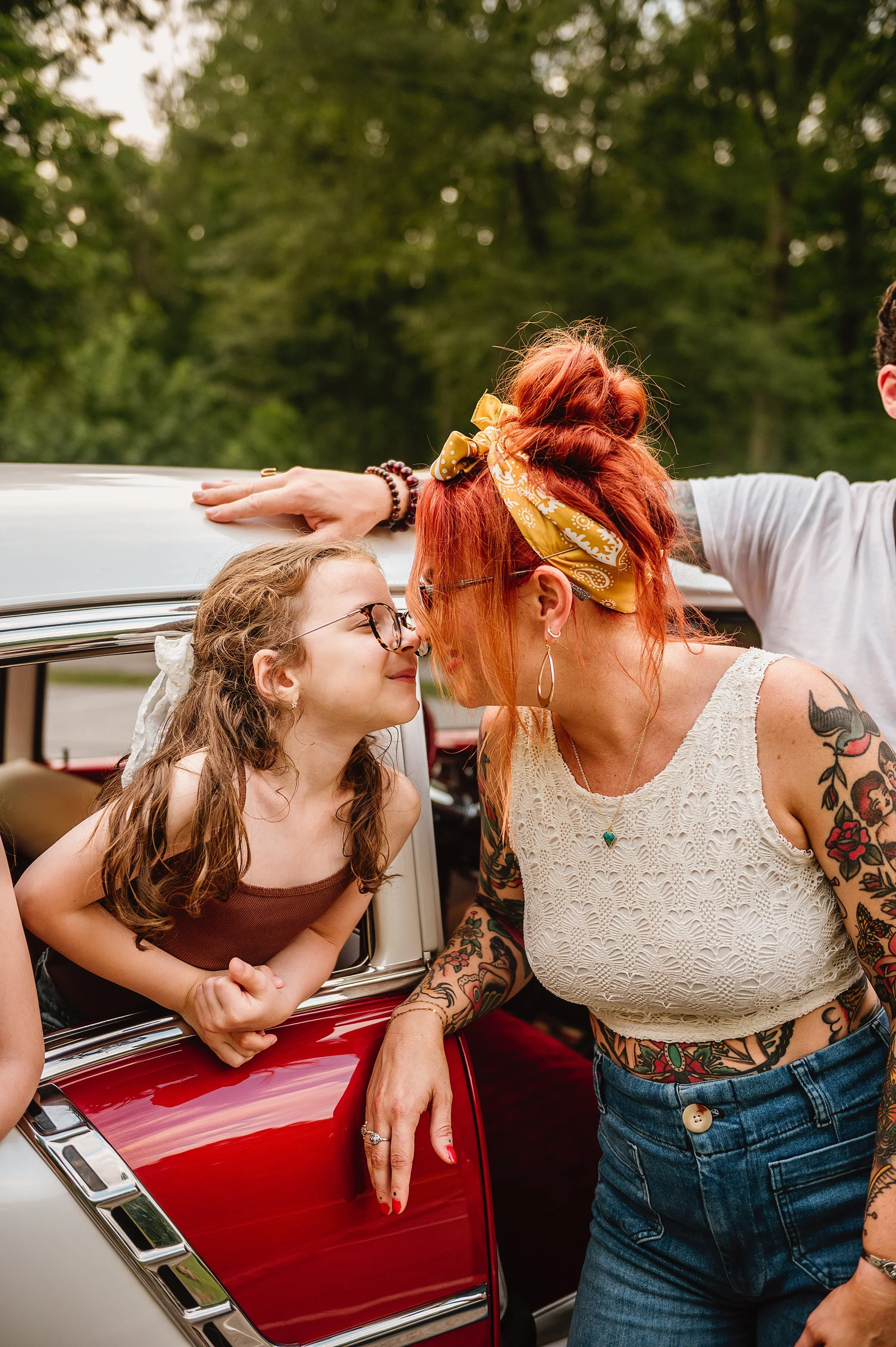 A woman with brightly colored hair and tattoos leaning on a red vintage car, sharing a moment with a young girl with glasses, in an outdoor setting with trees in the background.