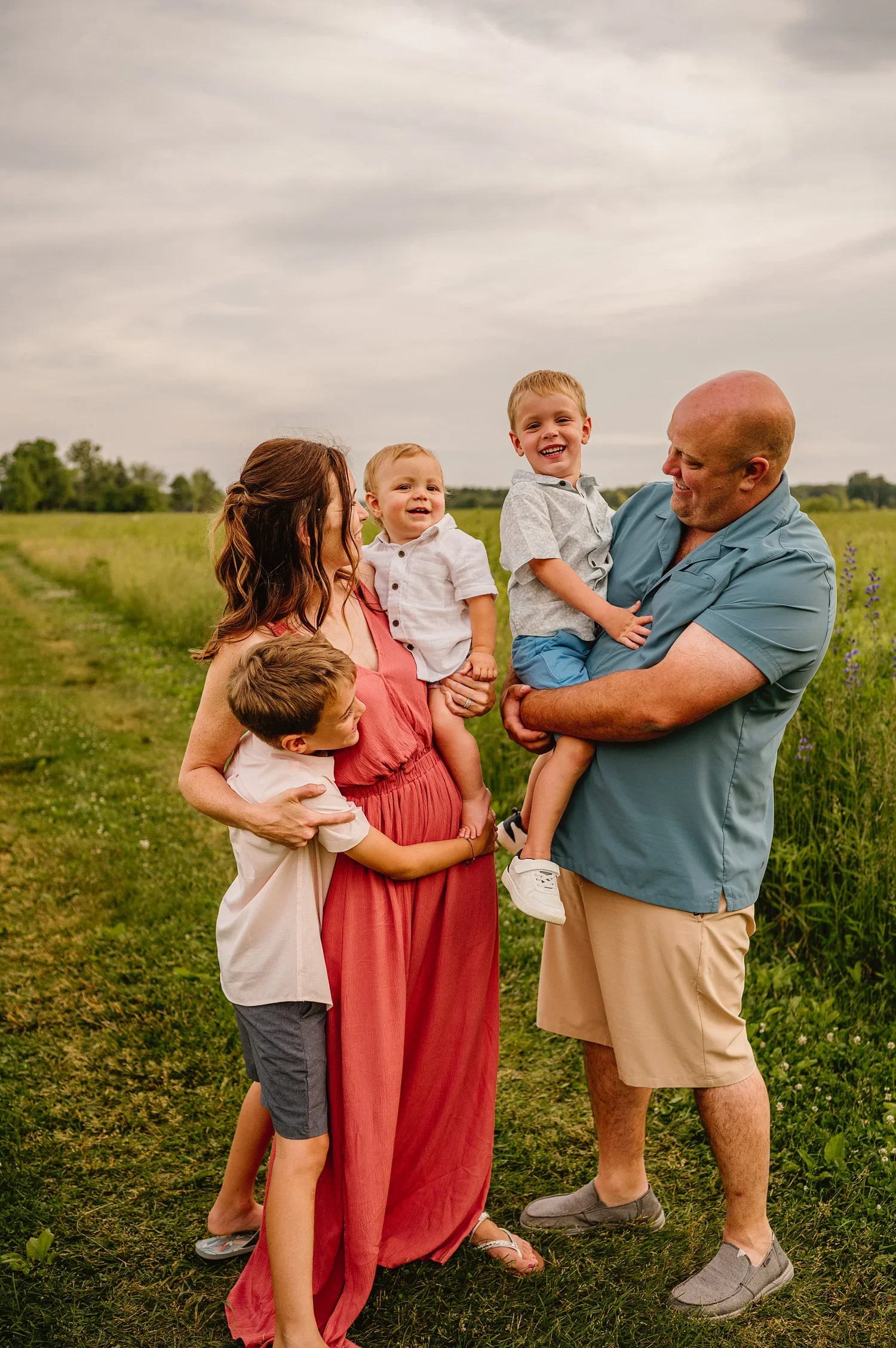 A family of six outdoors in a grassy field on a cloudy day, with three children, two women, and one man, all smiling and enjoying a moment together.