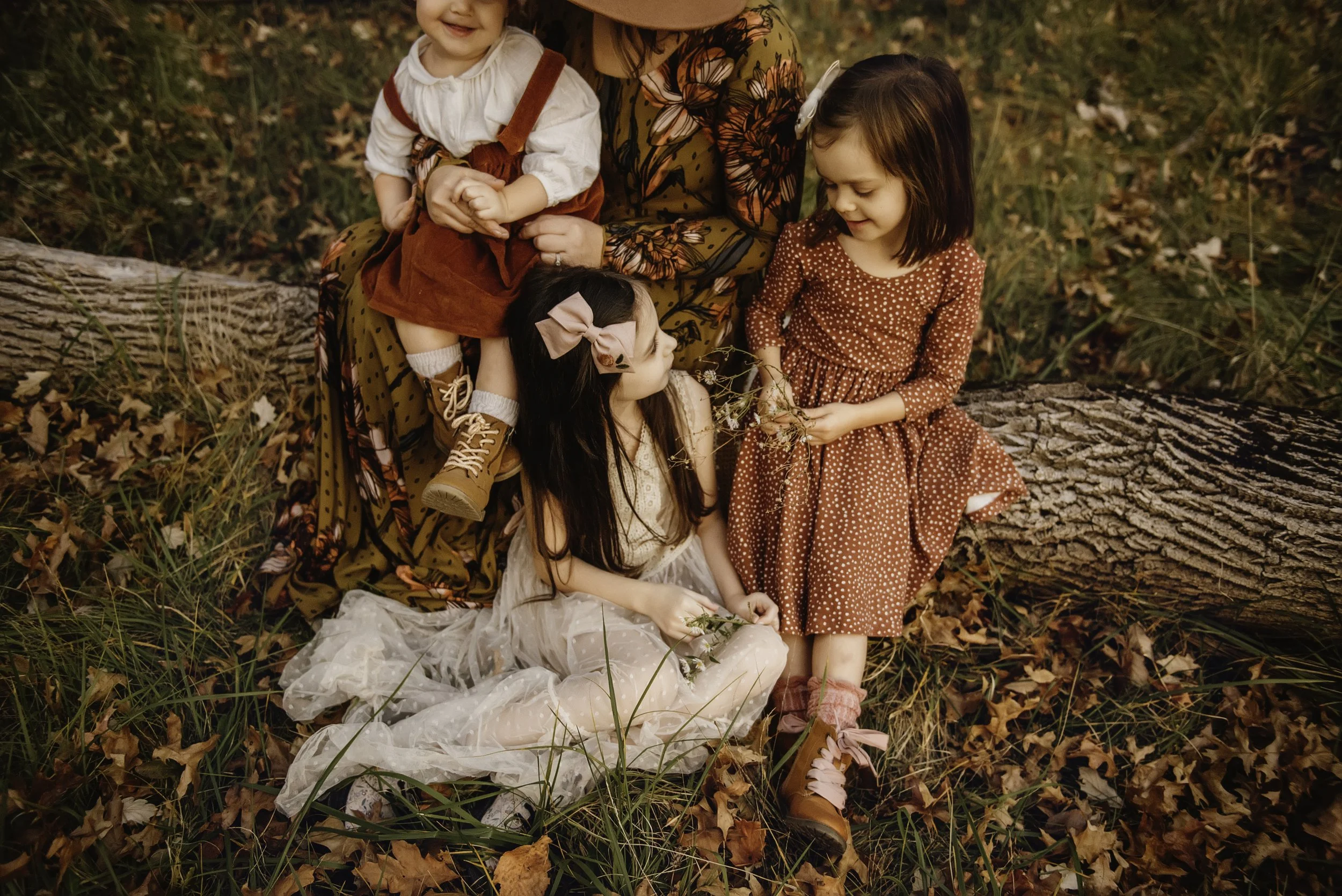 A woman with two young girls and a toddler sitting on grass and fallen leaves in autumn. The woman wears a floral dress and hat, one girl in a lace dress, the other in a polka dot dress, all wearing boots. The toddler is dressed in overalls.