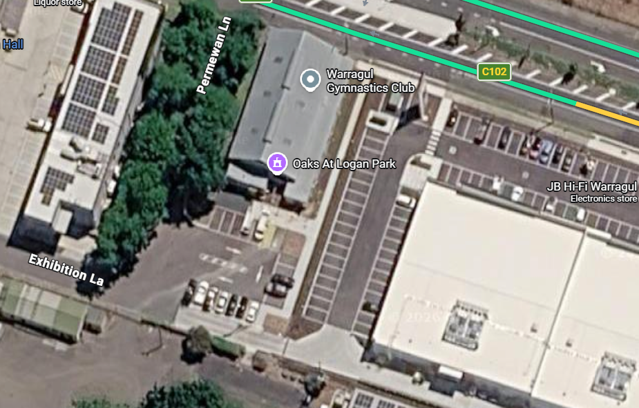 Satellite view of a commercial area showing parking lots, streets labeled 'Exhibition La' and 'Permanent Ln,' and buildings including Warragul Gymnastics Club, Oaks at Logan Park, and JB Hi-Fi Warragul electronics store.