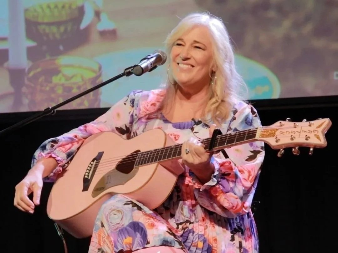 An older woman with long blonde hair playing an acoustic guitar while sitting on a stage, smiling, wearing a colorful floral dress. There is a microphone in front of her and a backdrop with blurred images.