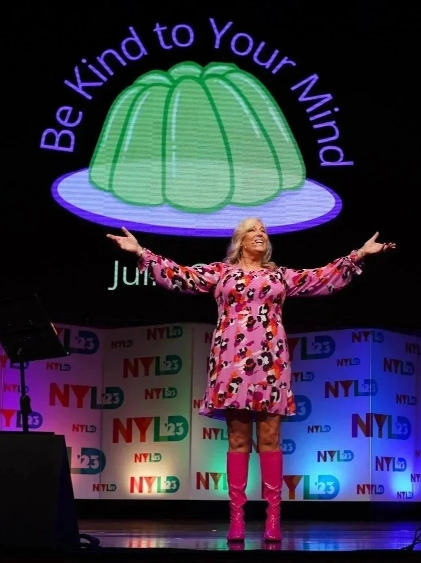 Woman standing on stage with arms outstretched, smiling, wearing a colorful pink and purple dress with matching pink boots, in front of a backdrop with NYL 23 logos and a large image of a green jelly. The large image has the text 'Be Kind to Your Mind' around it.