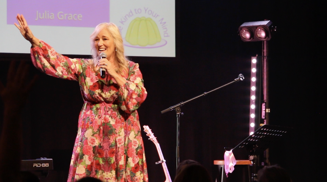 A woman with blonde hair, wearing a floral dress, speaking into a microphone on stage. There is a screen behind her with text and a logo, and stage lights are visible.