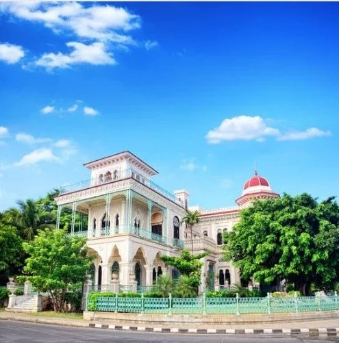 A grand mansion with intricate architecture and a red dome, surrounded by lush greenery, under a blue sky with white clouds.